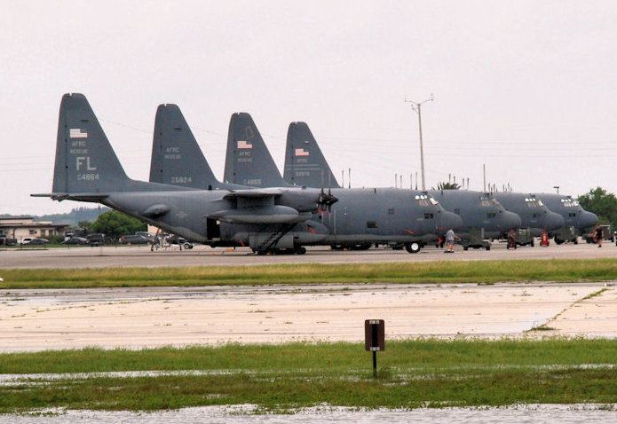 Four military cargo planes parked in a row on an airfield, with grass and runway in the foreground.