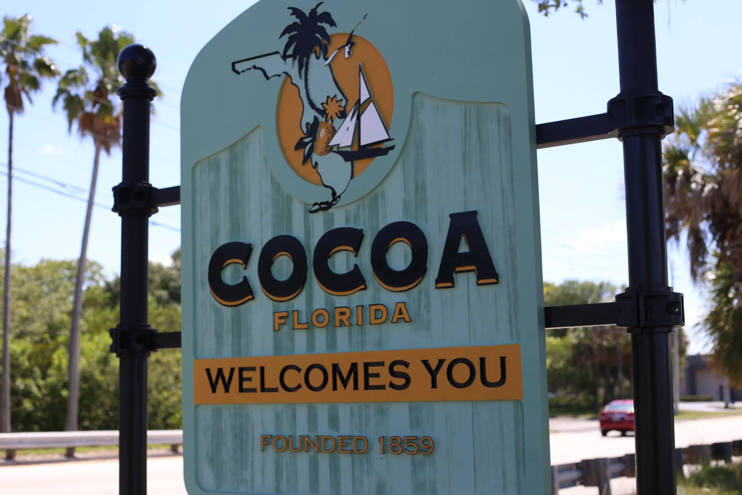 A welcome sign for Cocoa, Florida, showing a sailboat, palm trees, and the state of Florida outline, with a message welcoming visitors.