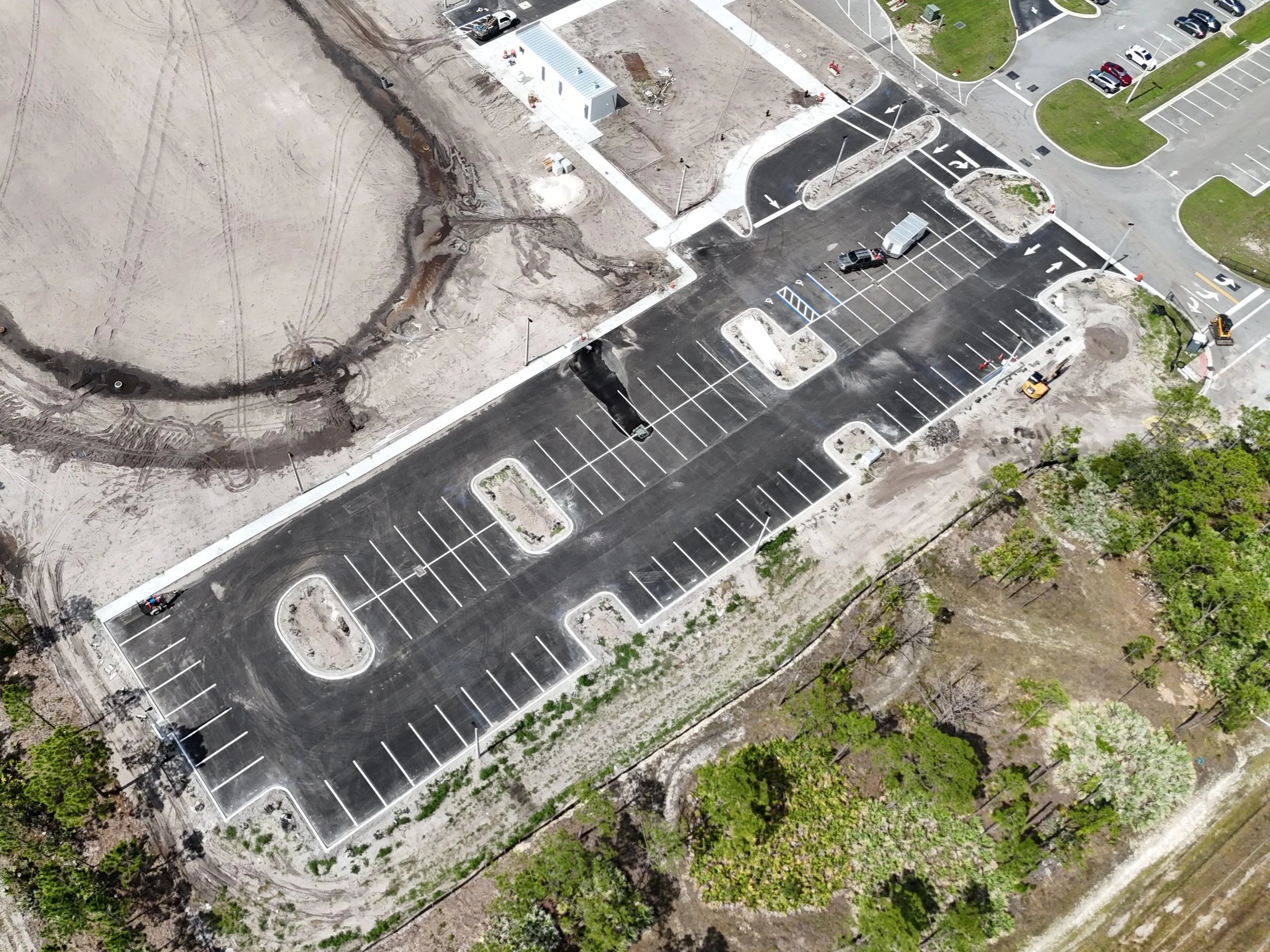 An aerial view of a parking lot with painted parking spaces and a few vehicles. Adjacent to the parking lot is a construction area with dirt, construction equipment, and signs of ongoing work.
