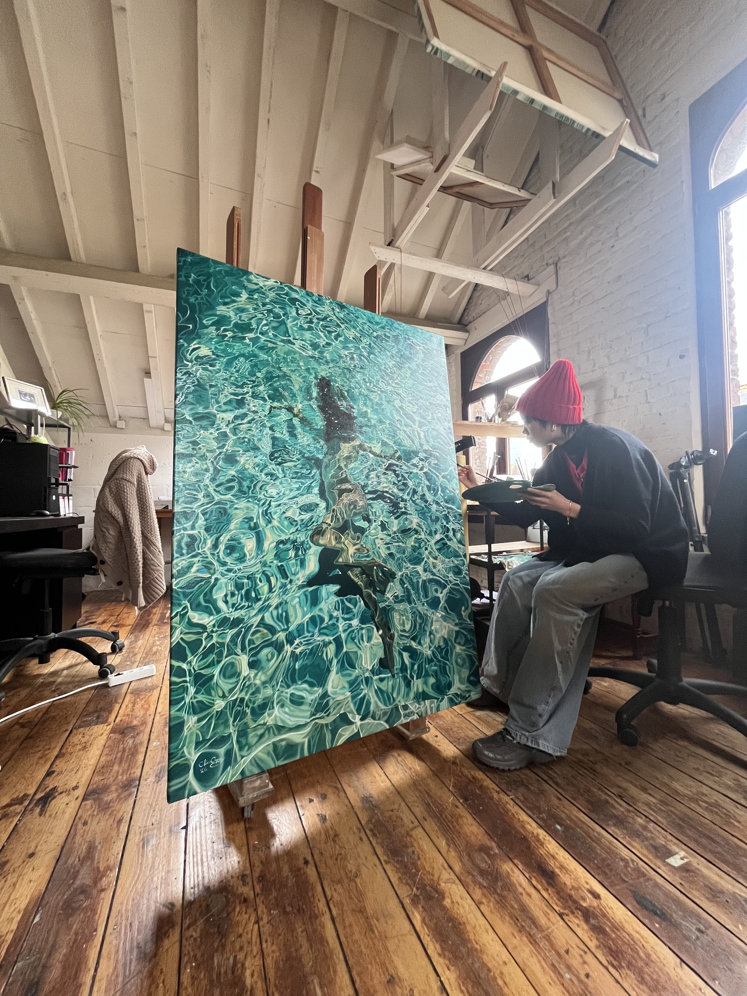 An artist painting a large water-themed artwork in a studio with wooden floors, white brick walls, and a person wearing a red beanie sitting and working with a palette.