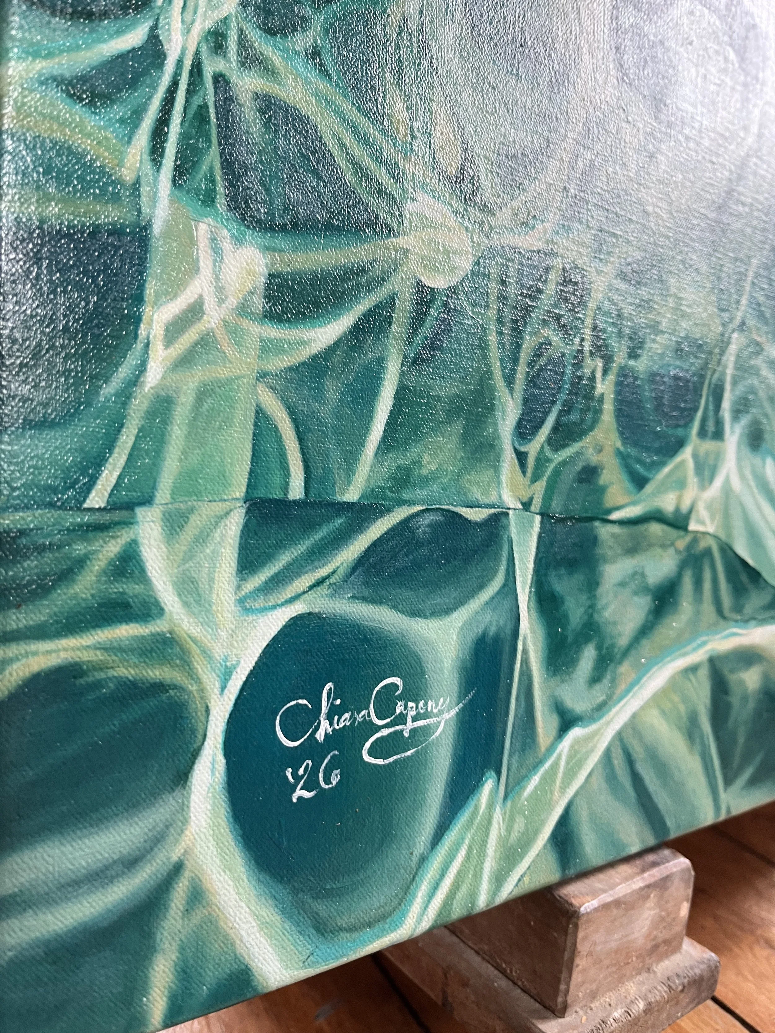 Close-up of a painting with shades of blue, green, and yellow, featuring white signature text and a price of '26'. The painting rests on two small bricks on a wooden surface.