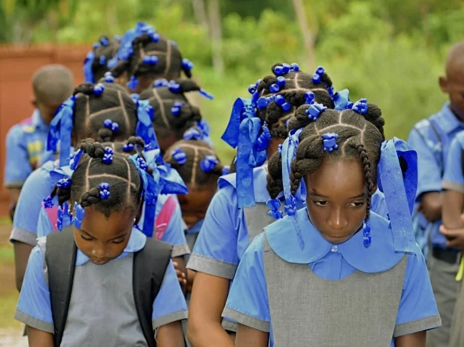 Young girls with braided hair decorated with blue ribbons and beads, wearing blue and gray school uniforms, standing outdoors.