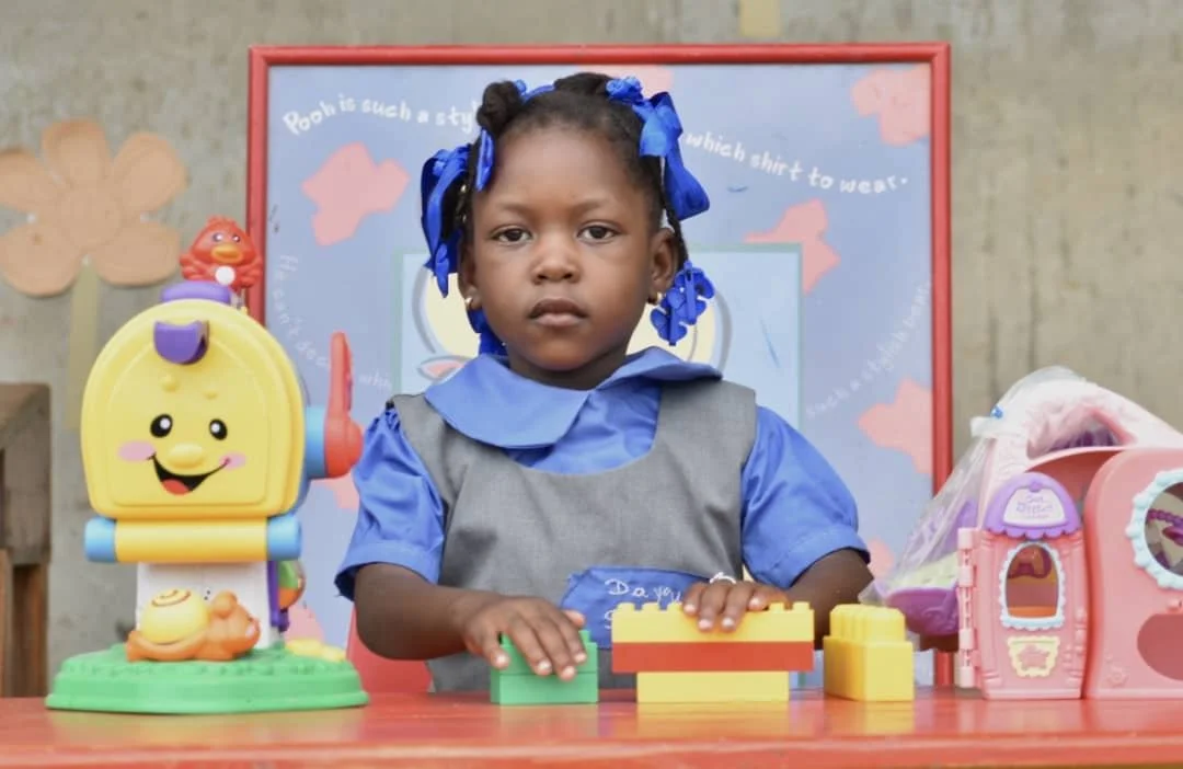 A young girl with braids and blue bows wearing a gray and blue school uniform, standing at a table with colorful toy blocks and toys, in front of a Pocoyo-themed classroom poster.