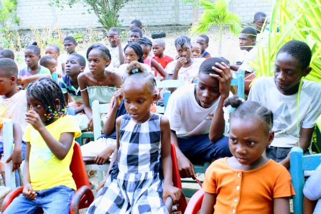 Group of children sitting outdoors on colorful chairs, some appear focused while others are distracted, with plants and a brick wall in the background.