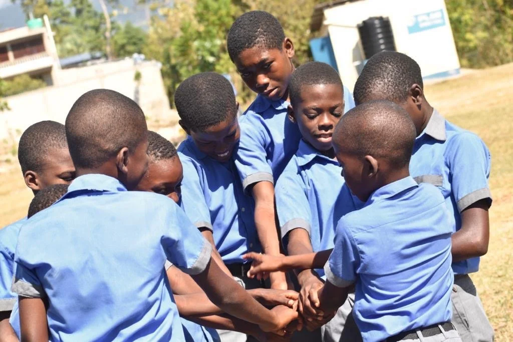 Group of young boys in blue school uniforms huddled together outside, doing a team huddle.