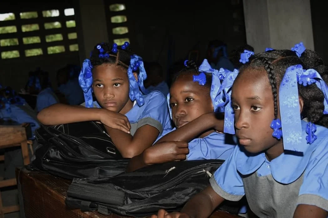 Three young girls in blue school uniforms with blue bows in their hair, sitting at wooden desks in a classroom, appearing bored or disinterested.