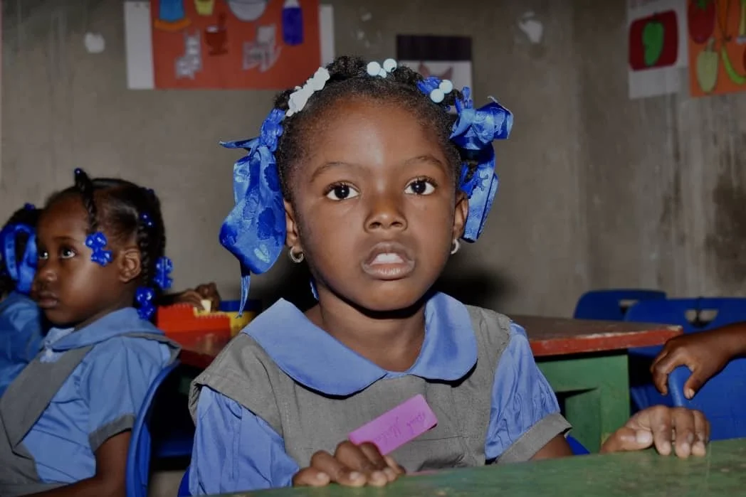 A young girl with braided hair decorated with blue ribbons and beads, wearing a school uniform, sitting at a desk in a classroom. There is another girl in similar attire in the background.