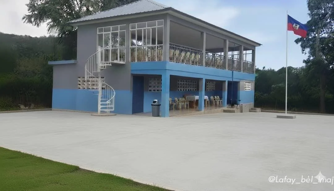 A two-story building with a white and blue exterior, an outdoor staircase on the left side, a covered balcony on the second floor, a flagpole with a flag, and several chairs and a table outside.