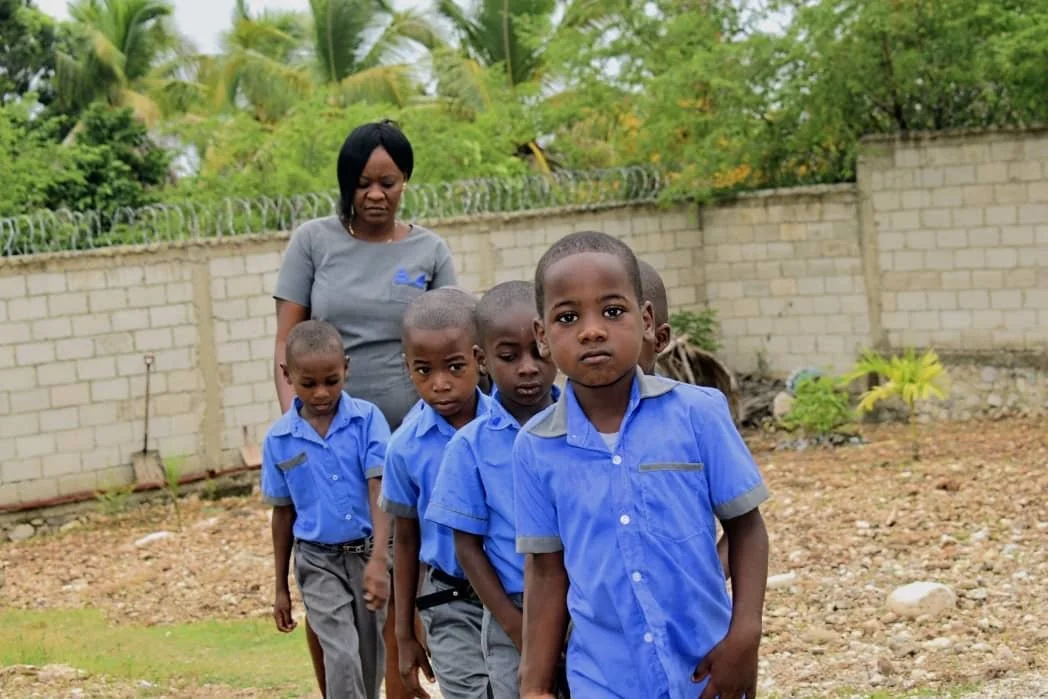 A woman and five young boys stand in a line outdoors near a brick wall, with green trees behind them. The boys are wearing matching blue shirts and gray pants, and the woman is wearing a gray shirt.