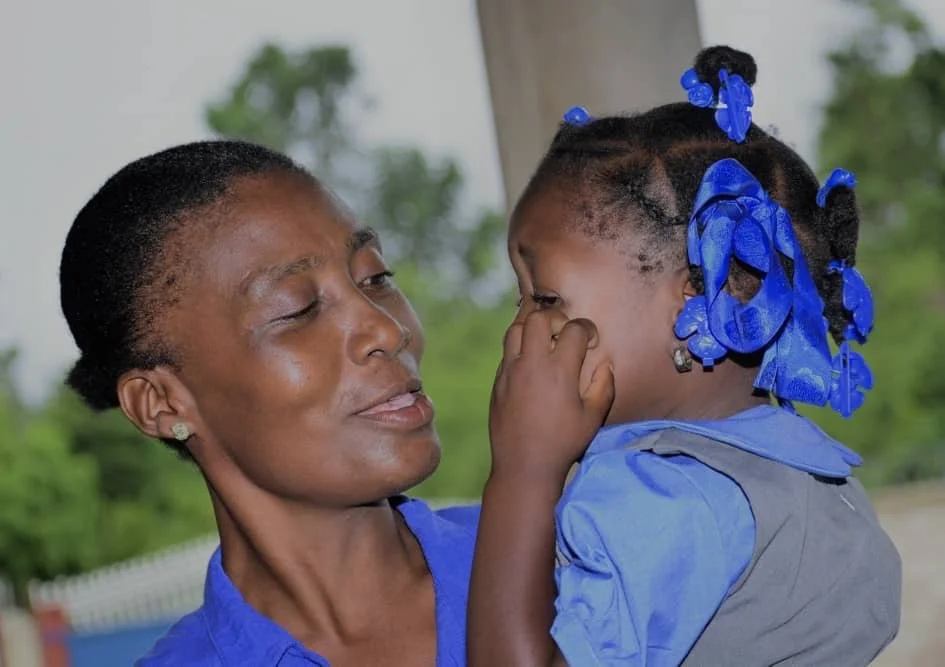 A woman and a young girl sharing a close moment, with the woman smiling and the girl covering her nose, both wearing blue clothing.
