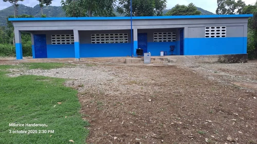 A single-story building painted in blue and gray with a flat roof, situated in a rural area with a dirt and gravel ground in front, greenery, and hills in the background.