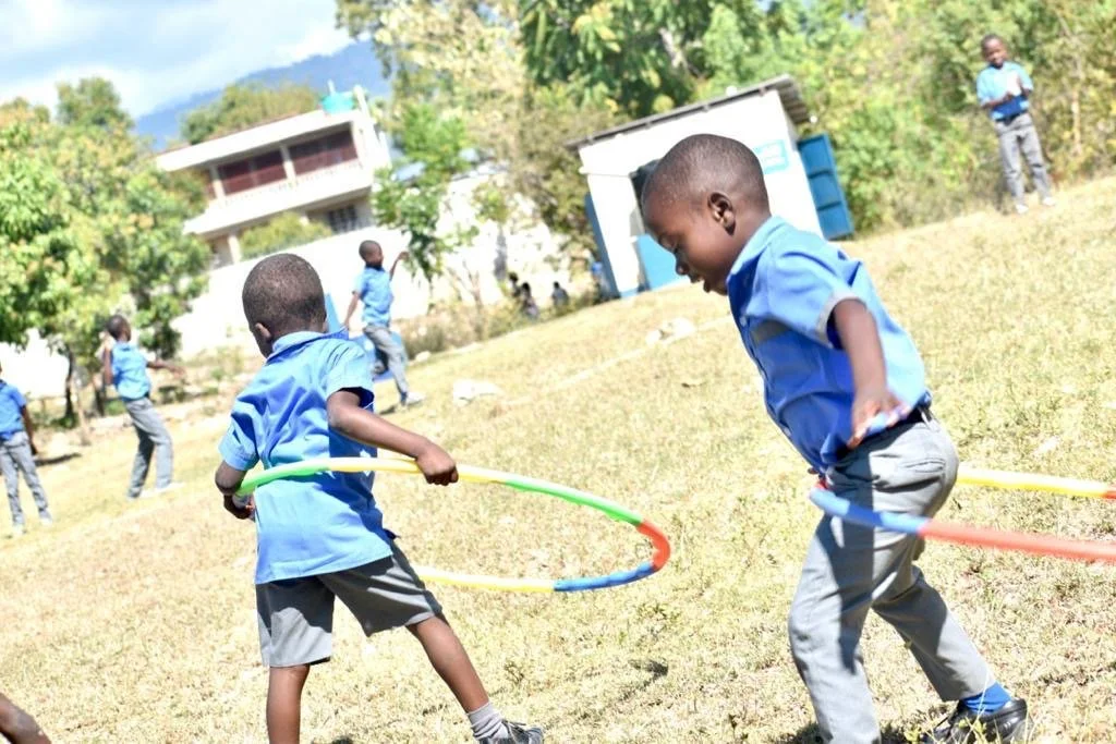 Children playing with hula hoops outdoors on a grassy field, with trees and buildings in the background.
