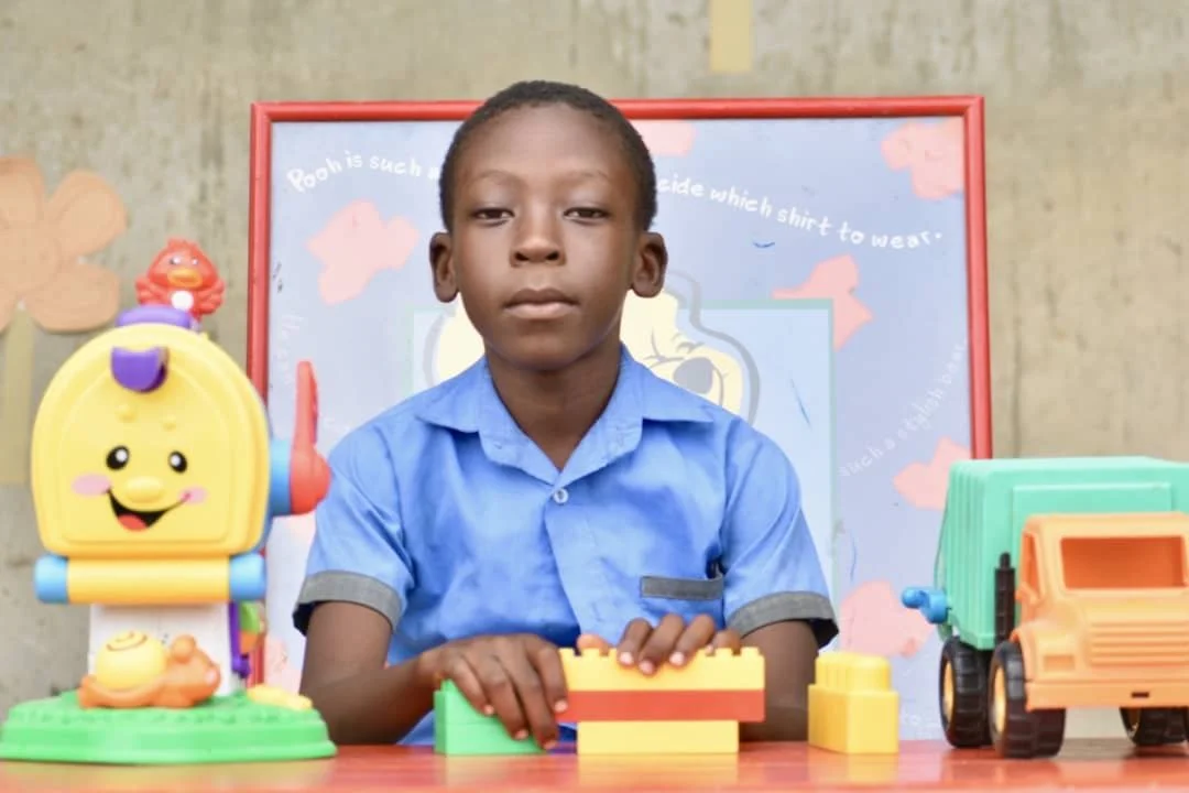 A boy in a blue shirt is sitting at a table with colorful toy blocks and toys, including a toy mailbox and a toy truck. There's a decorated background with a whiteboard and wall decorations.