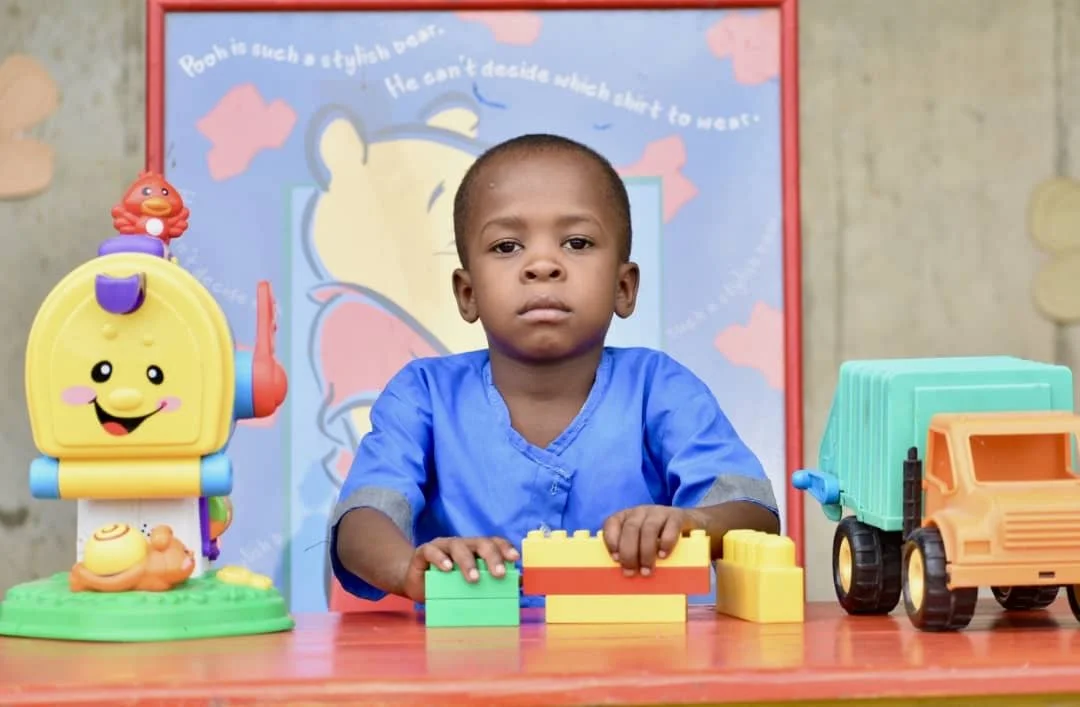 Young boy sitting at a table with colorful toy blocks, a toy truck, and a smiling colorful robot on the table. Background features a cartoon poster of Winnie the Pooh with a quote.