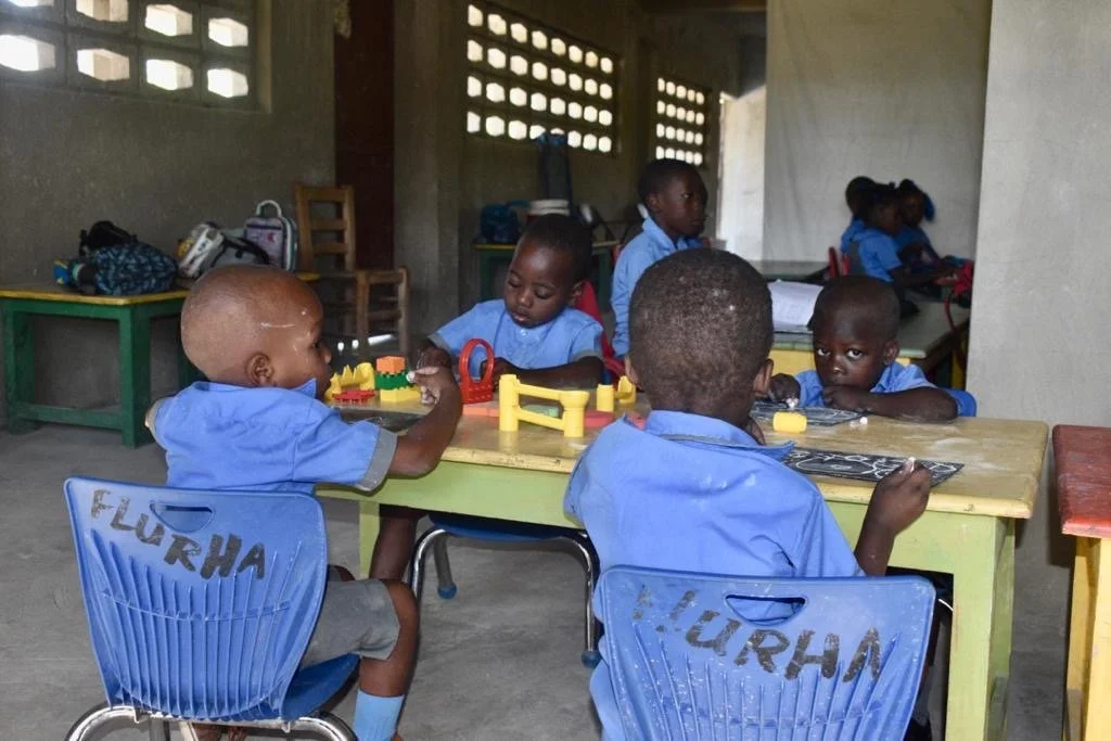 Young children in blue school uniforms sitting at tables, engaged in activities inside a classroom with concrete walls and small windows.