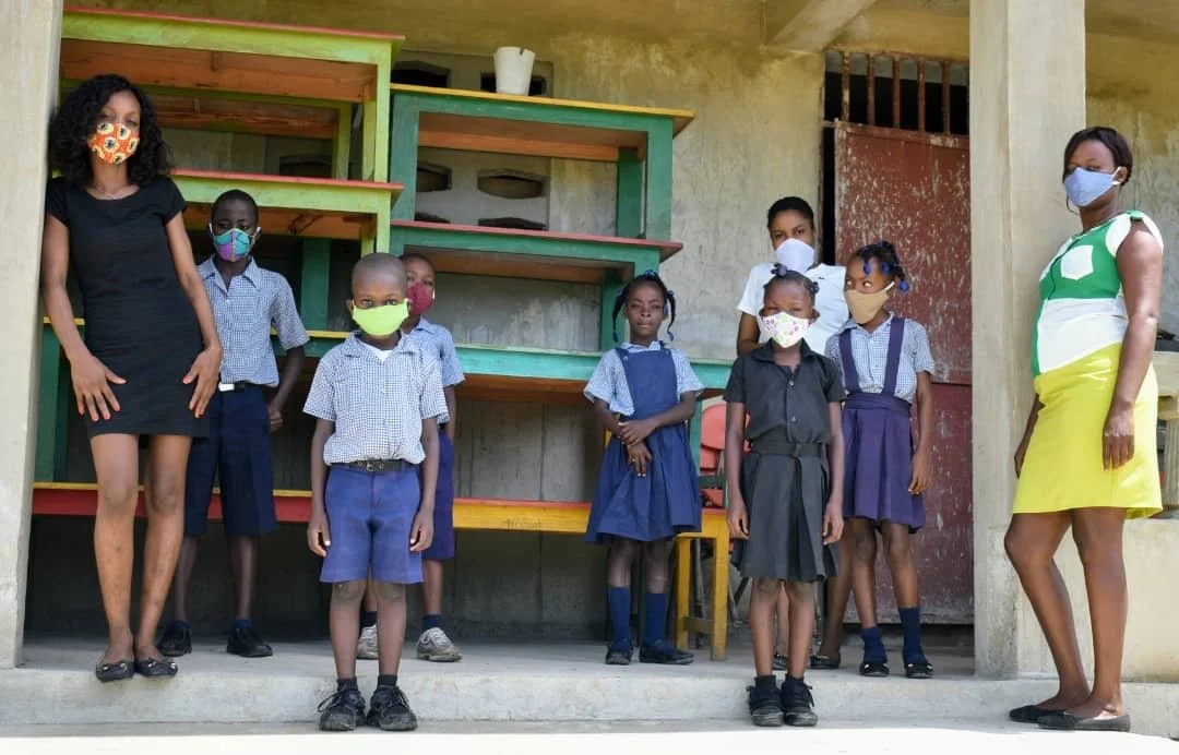 Group of children and two adult women wearing masks, standing outside a school classroom, some in school uniforms, with colorful shelves in the background.