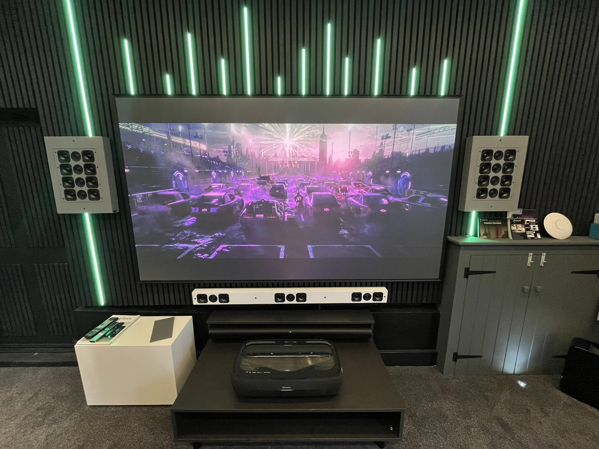Home theater setup with large screen displaying a futuristic cityscape, surround sound speakers, and a projector on a stand. LED strip lights enhance the ambiance.