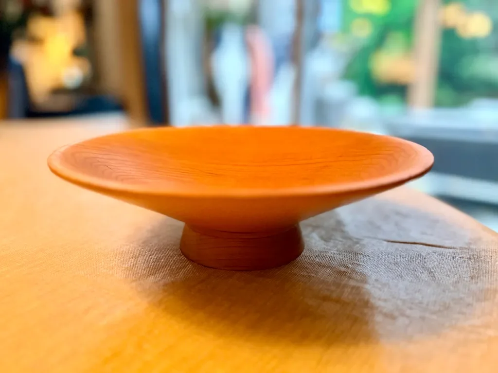 A small, wooden bowl made from cherry on a table near a window with a blurred outdoor view.