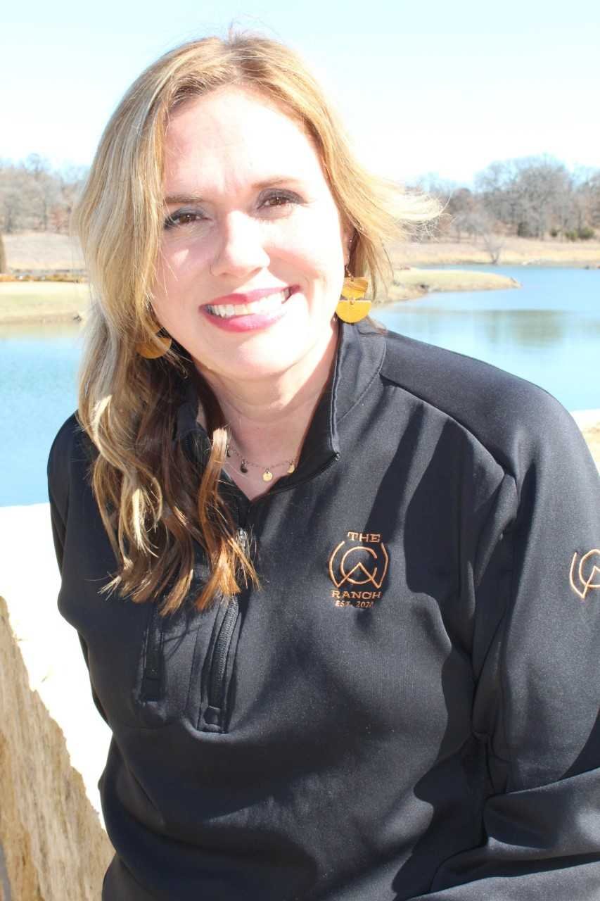 Woman in black jacket standing outside by a pond with trees in the background.