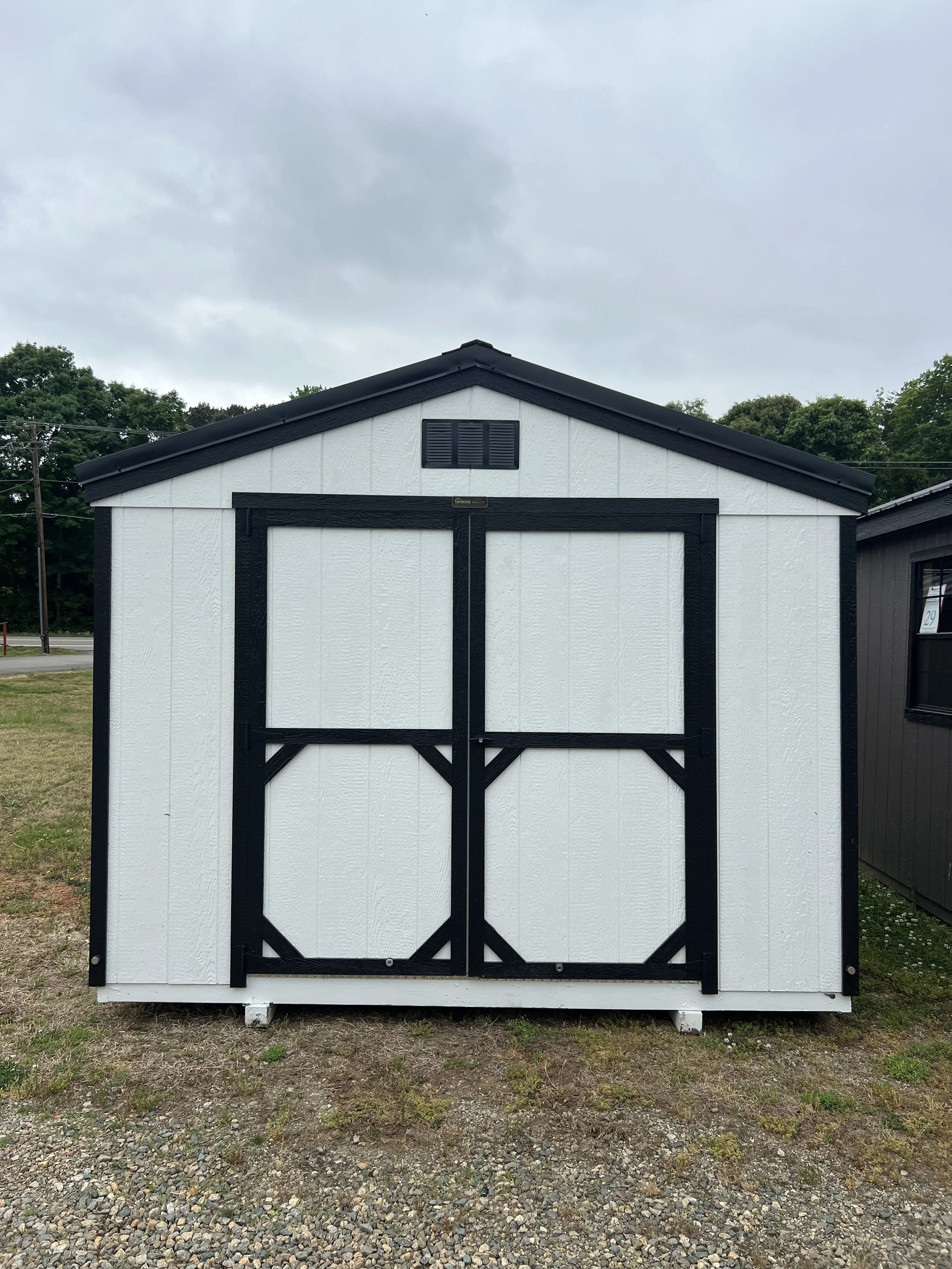 A white/black outdoor shed with a dark roof and double doors, located in a grassy area with trees in the background.