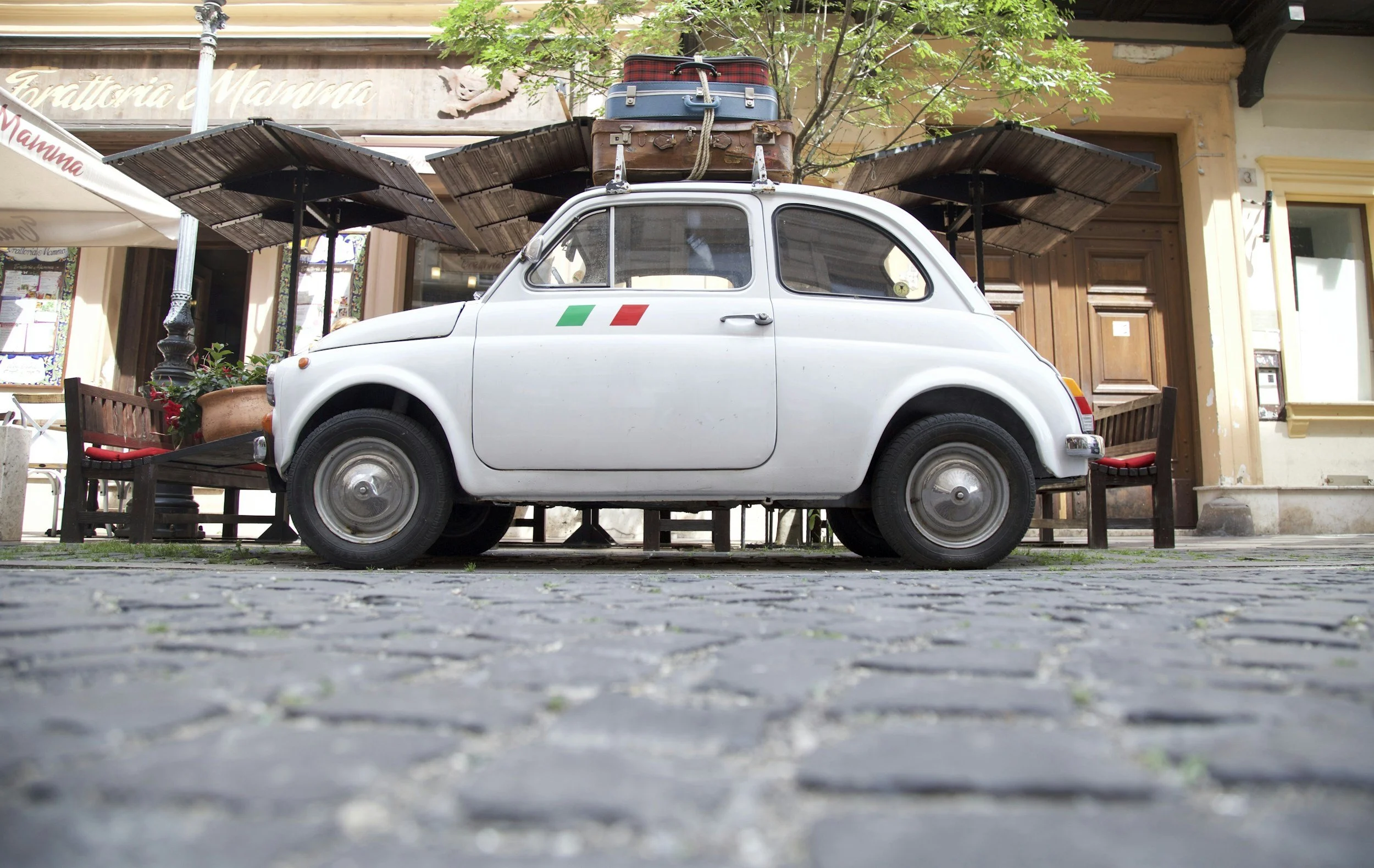 Een witte kleine auto met Italiaanse vlag op de deur, geparkeerd op een straat met huizen op de achtergrond, met bagage op het dak, waaronder koffers en tassen.