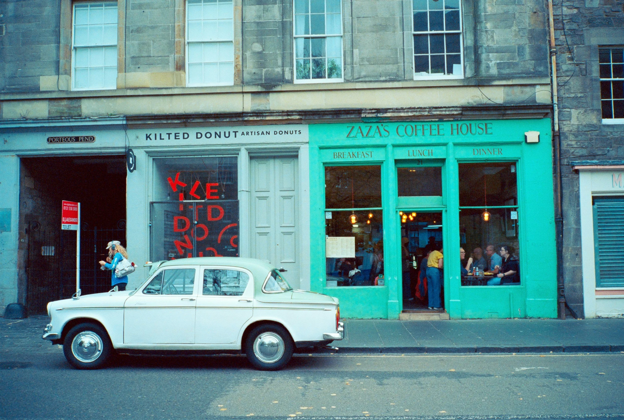Straatbeeld met een wit vintage auto en een horecagelegenheid met groen gevel en grote ramen, mensen zitten binnen, gevel heeft teksten 'Zaza's Coffee House' en 'Kilted Donut'.