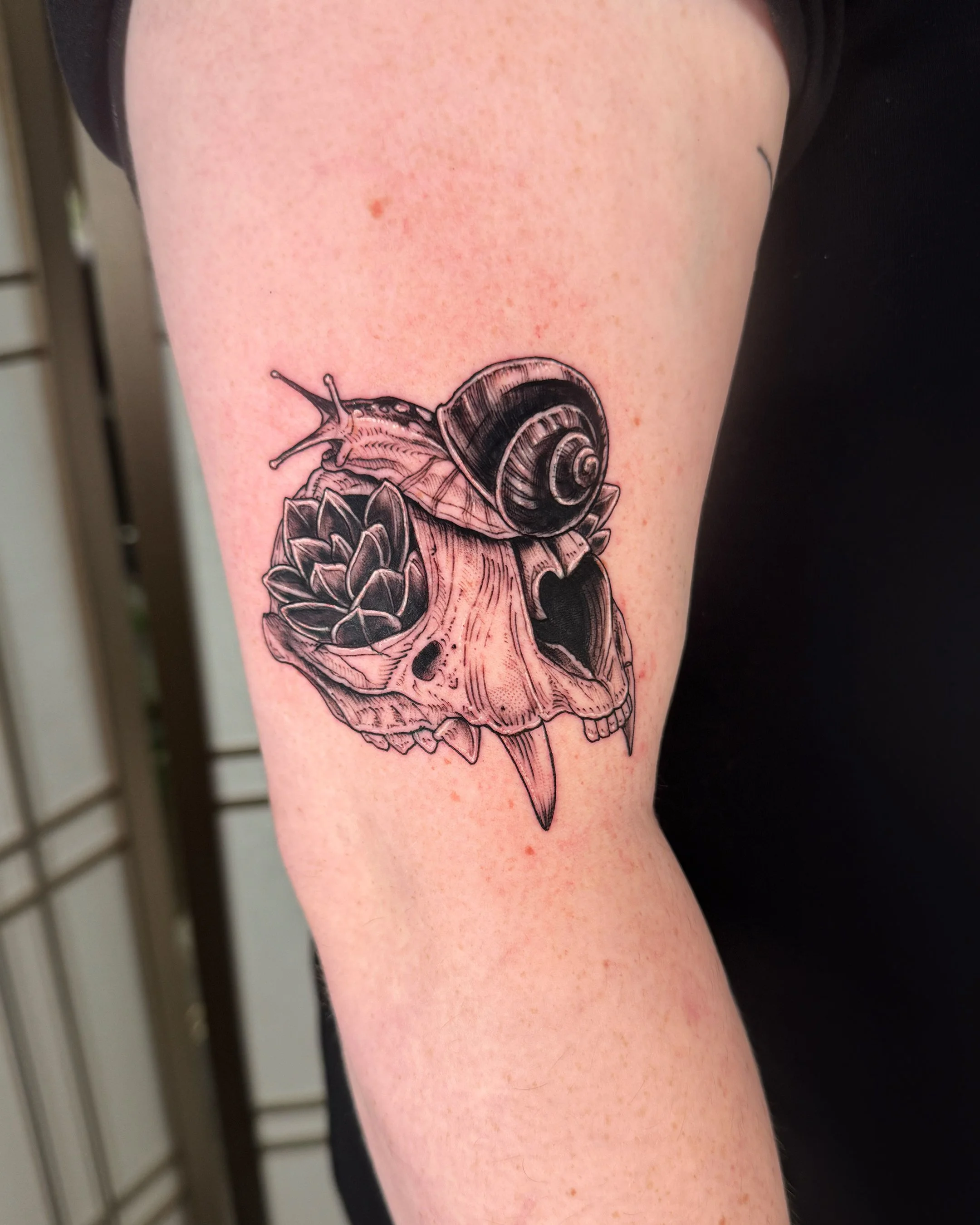Black and gray tattoo of a snail on a human arm, with a large shell and detailed body, positioned above a floral design.