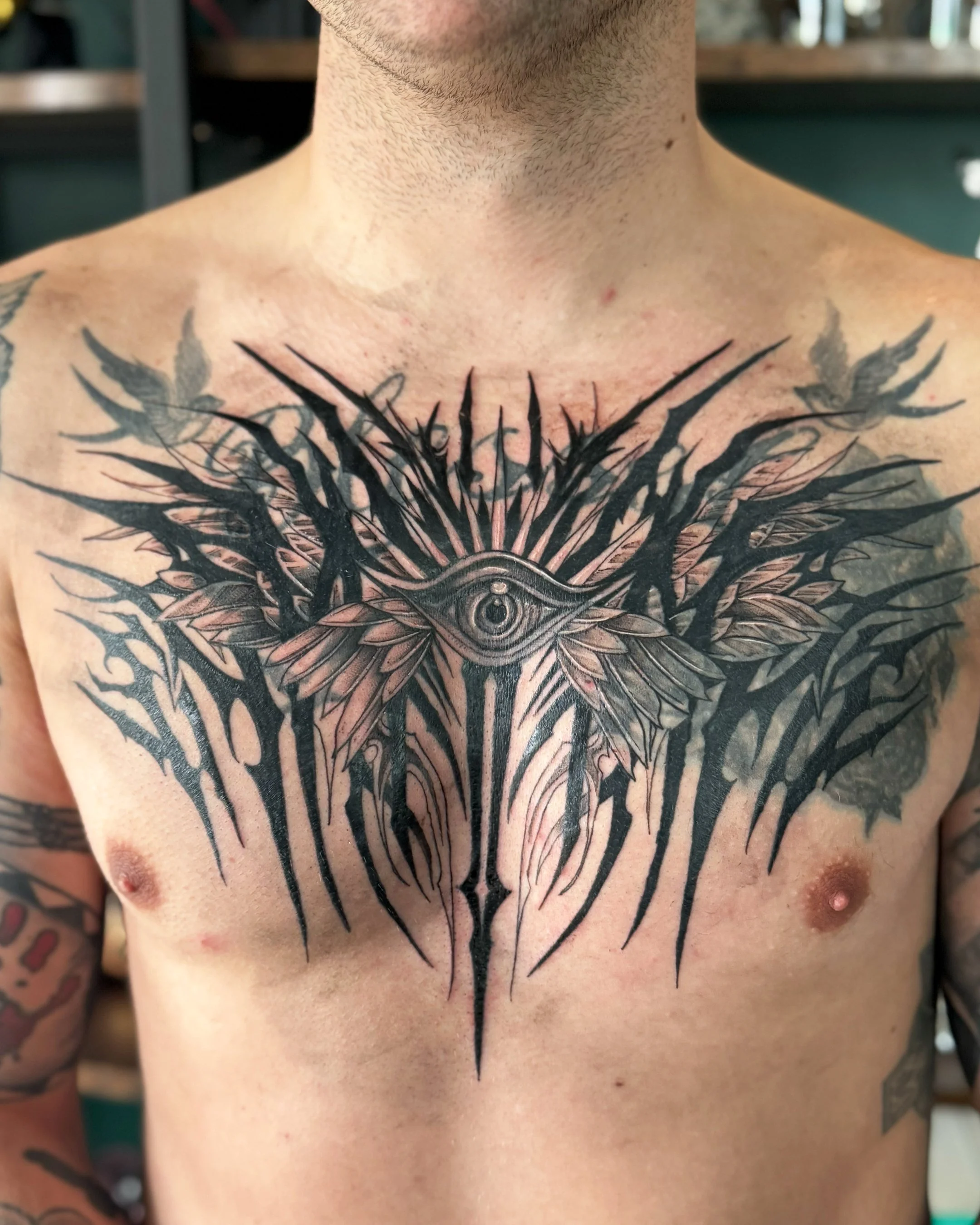 A tattoo of a detailed eye at the center with intricate black lines and patterns extending across the chest, with some visible tattoos on the person's arm.