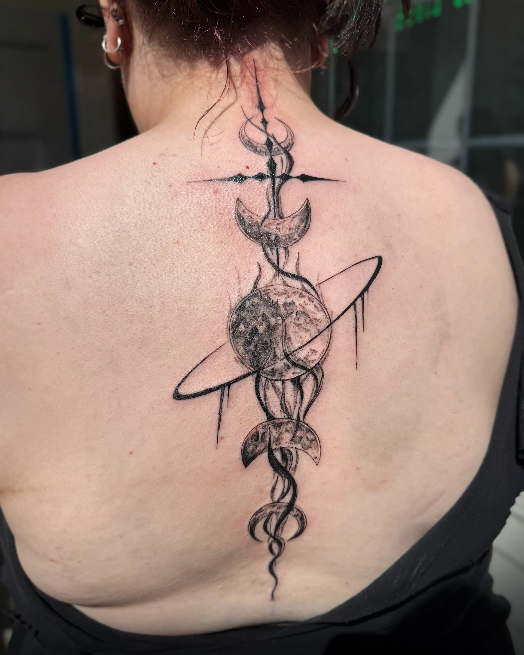 A tattoo on a person's upper back and neck, featuring a design that includes a detailed moon connected to a star and other celestial elements, drawn in black ink.