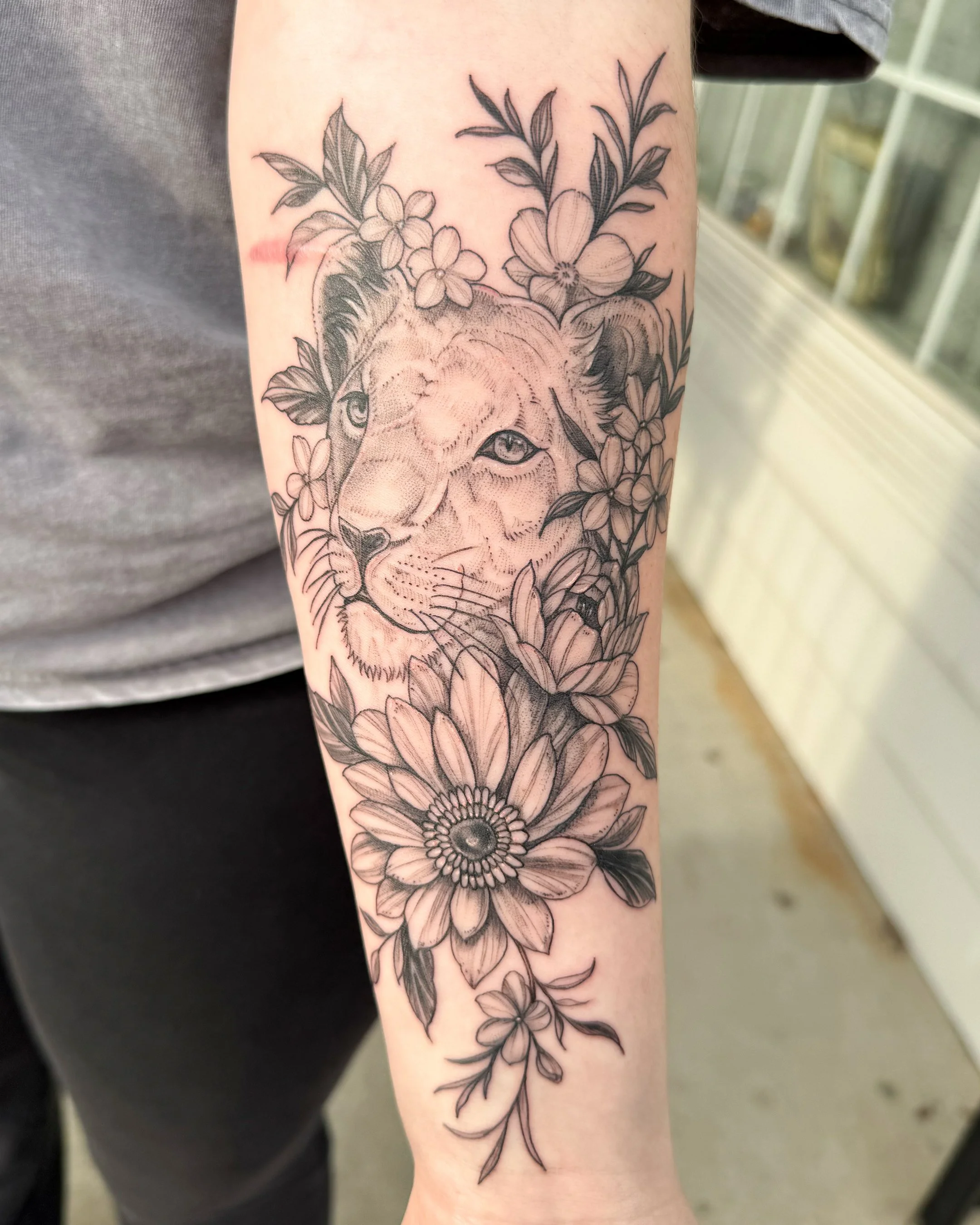 Black and gray floral tattoo on a person's arm, featuring a lion's face surrounded by flowers and leaves.