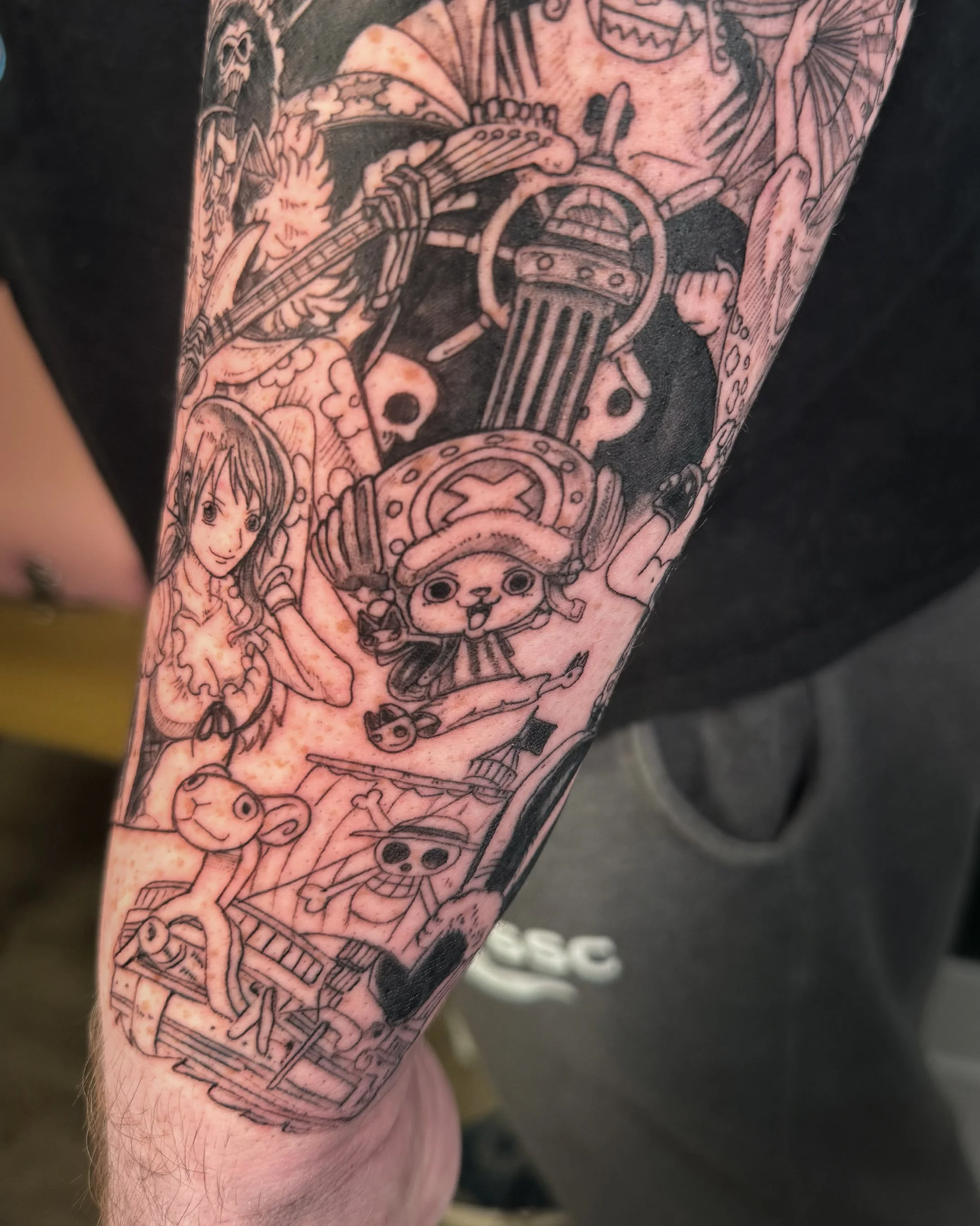 A tattoo sleeve featuring various characters and objects, including a girl with long hair, a smiling skull, a skull with crossbones, a ghost, and other detailed images.