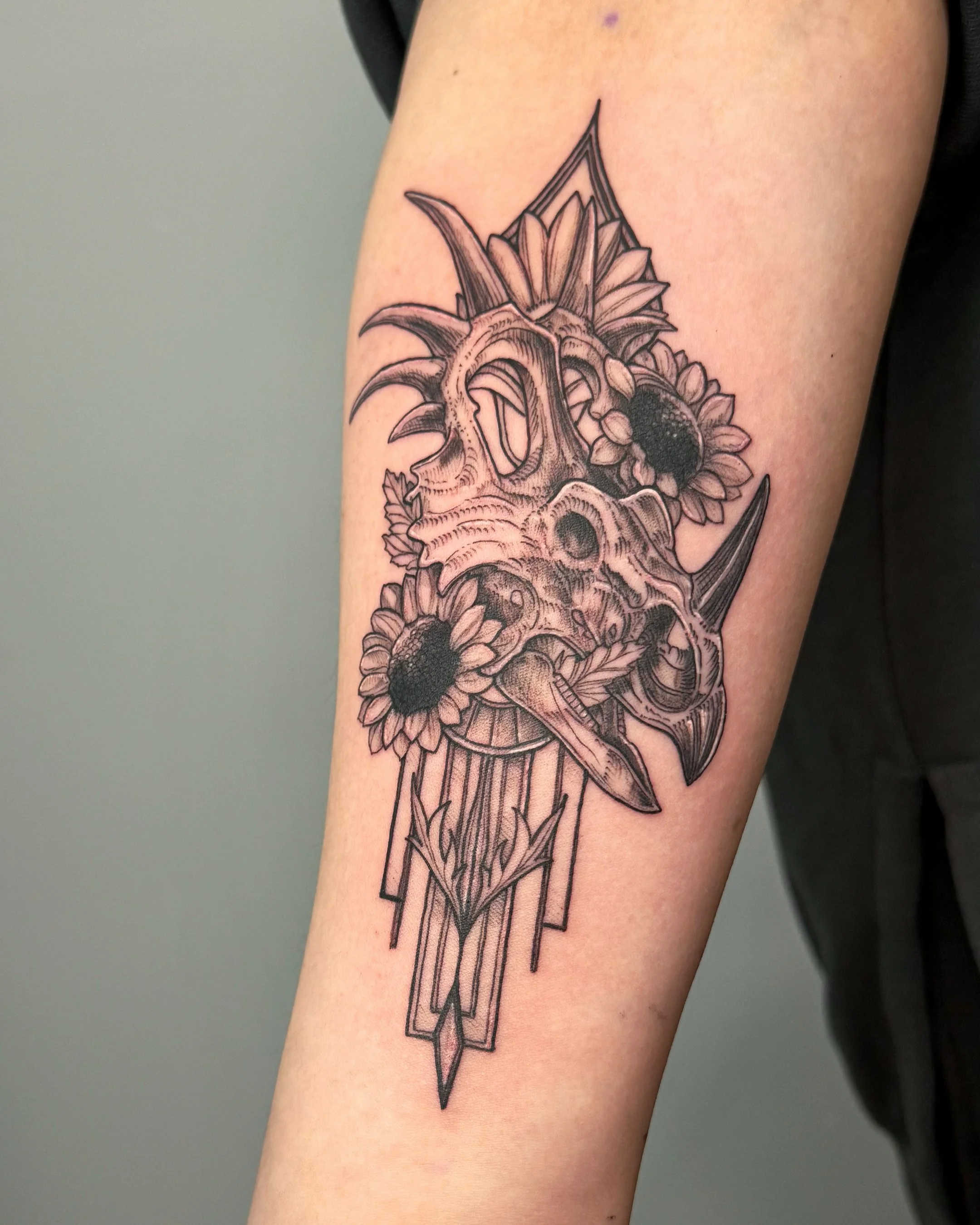 Black and gray tattoo of a dragon skull with flowers and geometric shapes on a person's upper arm.