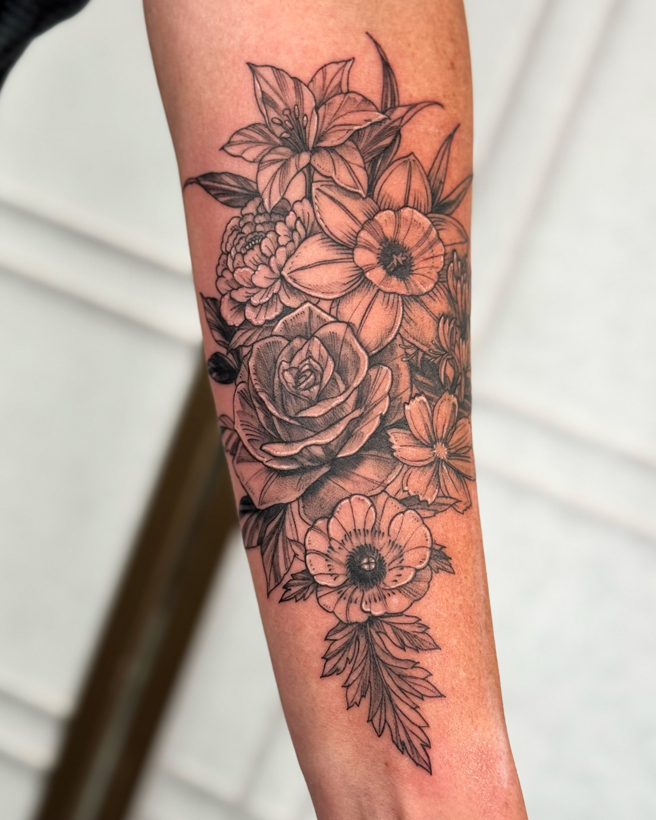 A black and gray floral tattoo on a person's forearm, featuring various flowers including a lily, anemone, and rose, with leaves and detailed linework.