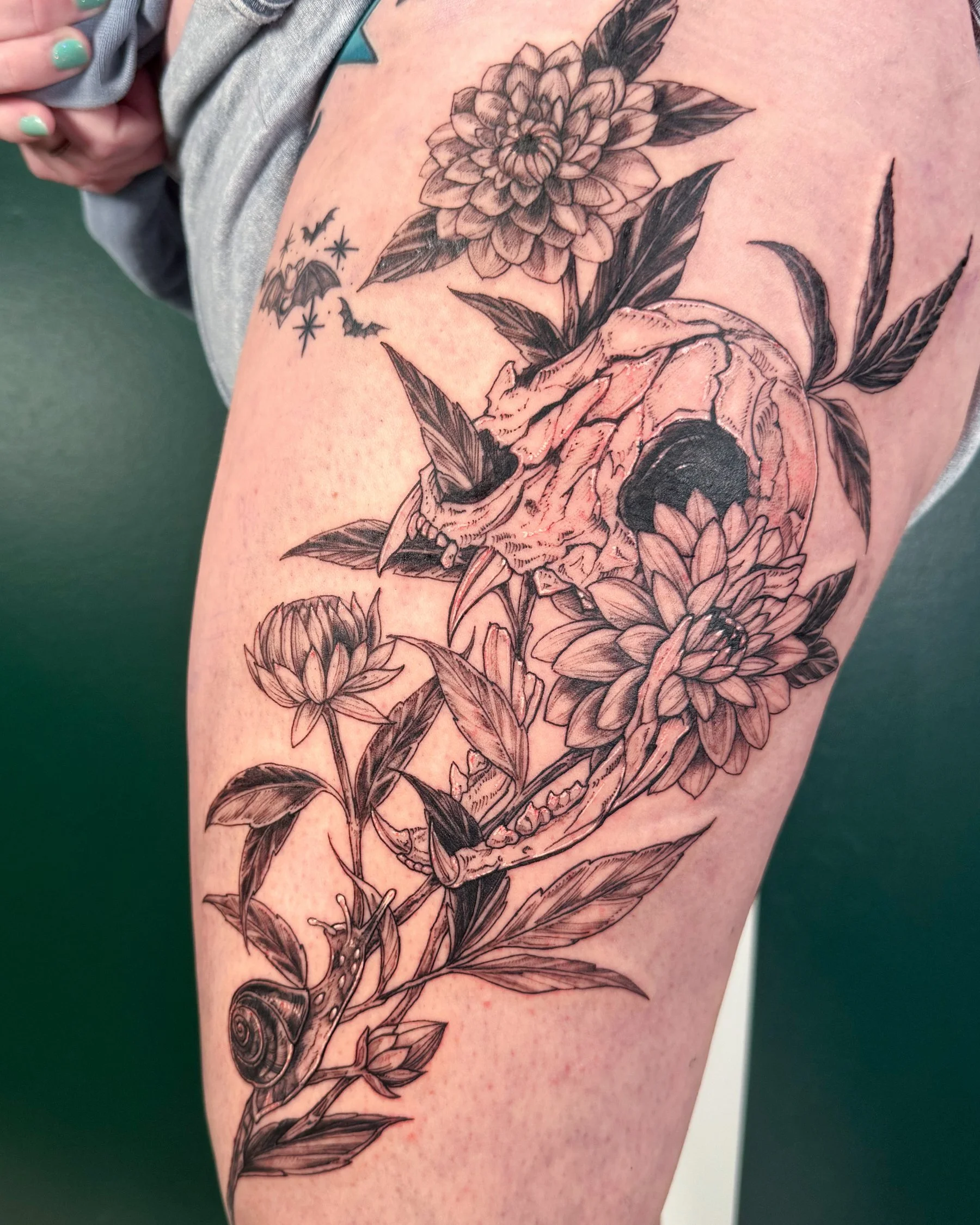 A black and gray tattoo of a skull with floral and botanical elements, including a large flower, leaves, bats, stars, and a snail shell, on a person's upper arm or shoulder.