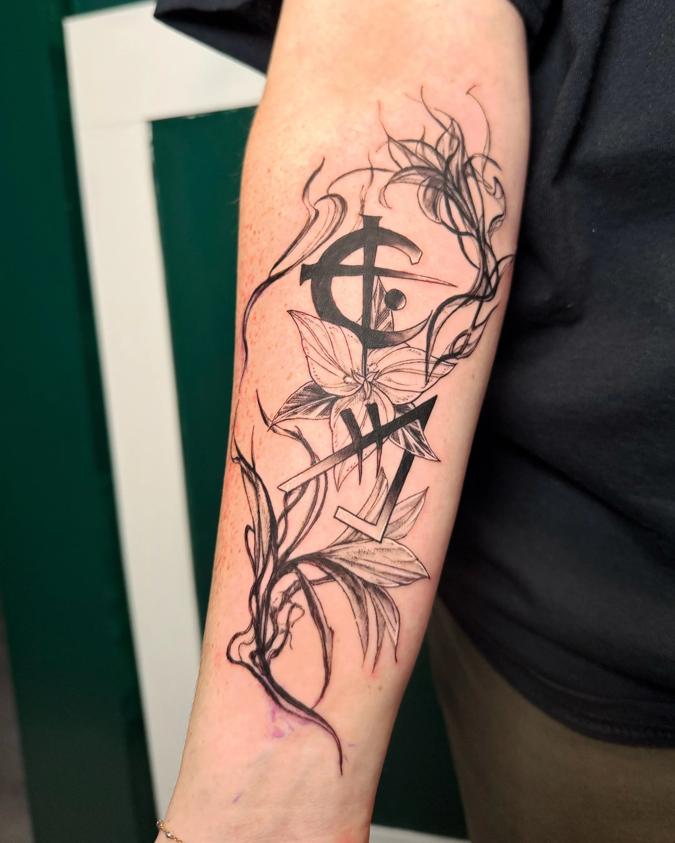 A fresh tattoo on a person's arm featuring black and grey floral design with detailed leaves and flowers. The tattoo appears to be in the process of healing or recently done.