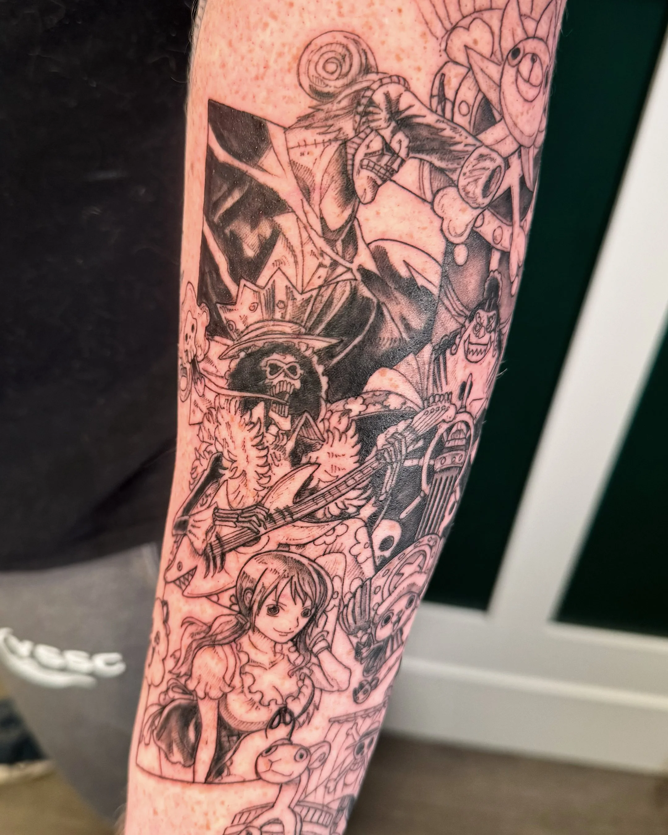 A tattoo on a person's forearm featuring various sketches of characters and elements from Japanese manga and anime, including a girl with long hair, a skull, and a mechanical object, all in black ink with detailed shading.