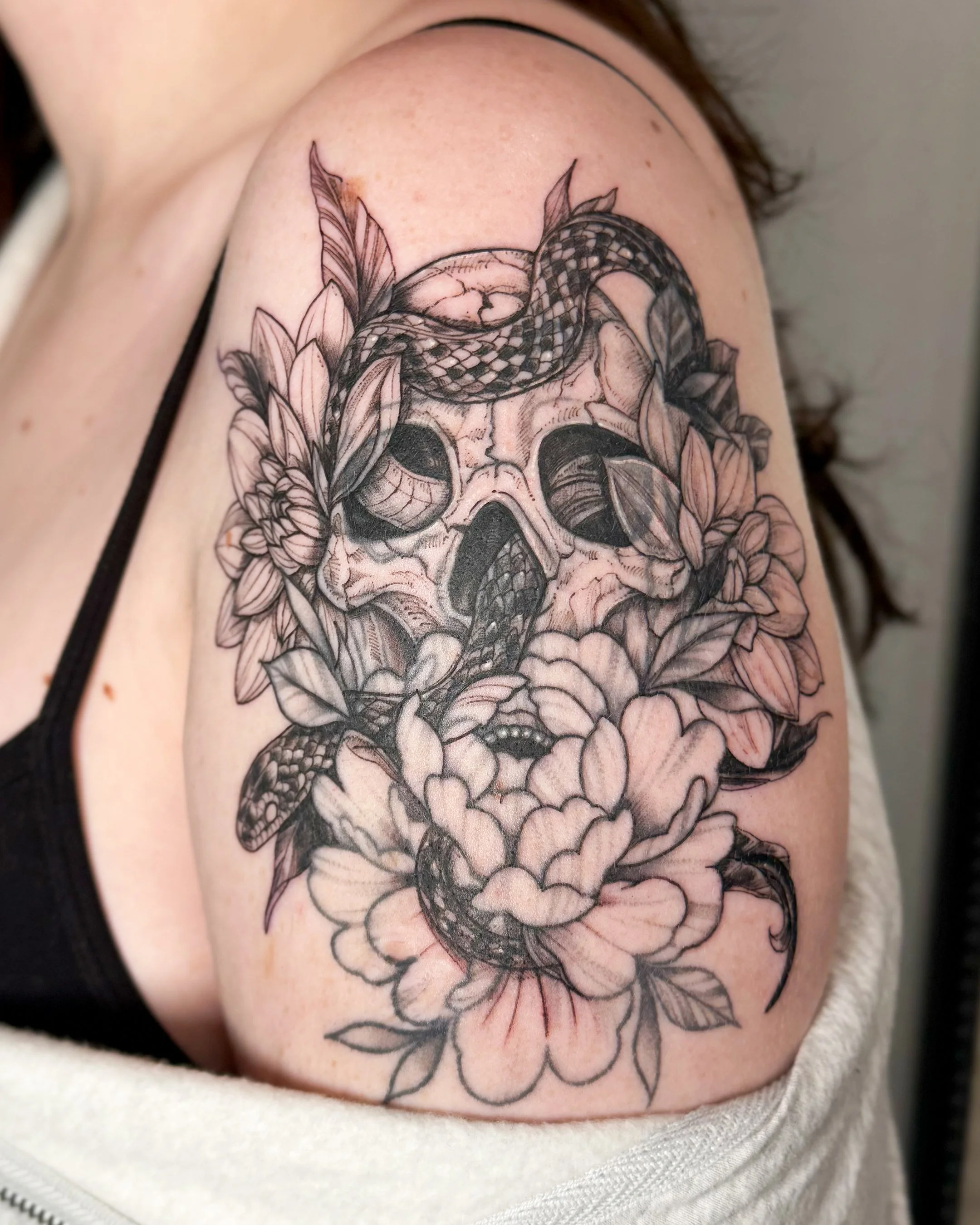 A black and gray tattoo of a skull with a snake wrapped around it, surrounded by flowers on someone's upper arm.