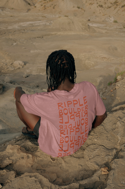 Man sitting outside on dirt and rocks wearing ripple boulder shirt with back of shirt facing camera. The design repeats the Ripple Boulder logo in a decorative way down the back of the shirt with pink cotton cloth and red text.