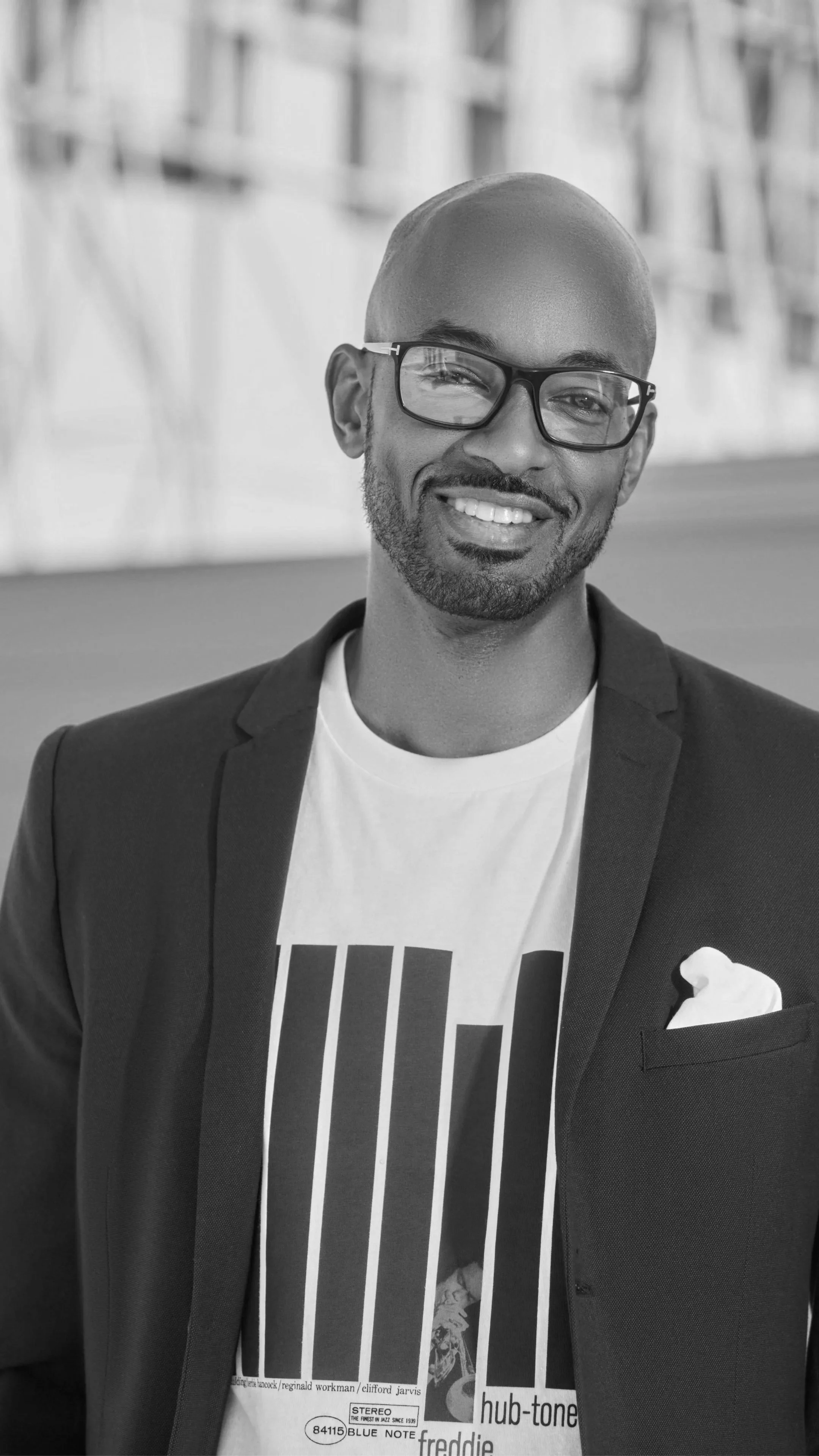 Black and white photo of a smiling man wearing glasses and a suit jacket, standing in front of a blurred background.