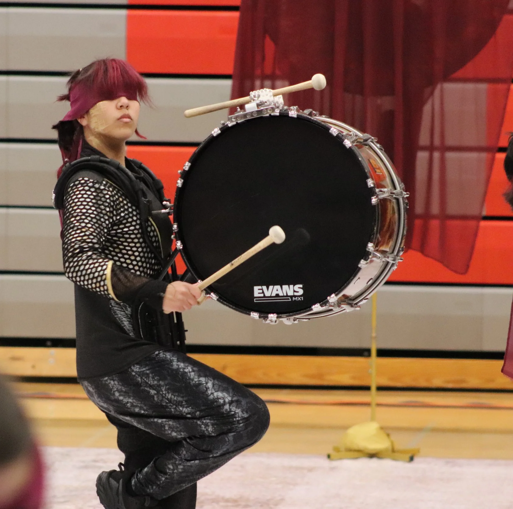 A young person dressed in black with a fishnet-style shirt and patterned pants, holding drumsticks, blindfolded, playing a large bass drum labeled 'EVANS' on a gymnasium gym floor. Indoor Percussion drumline bass player