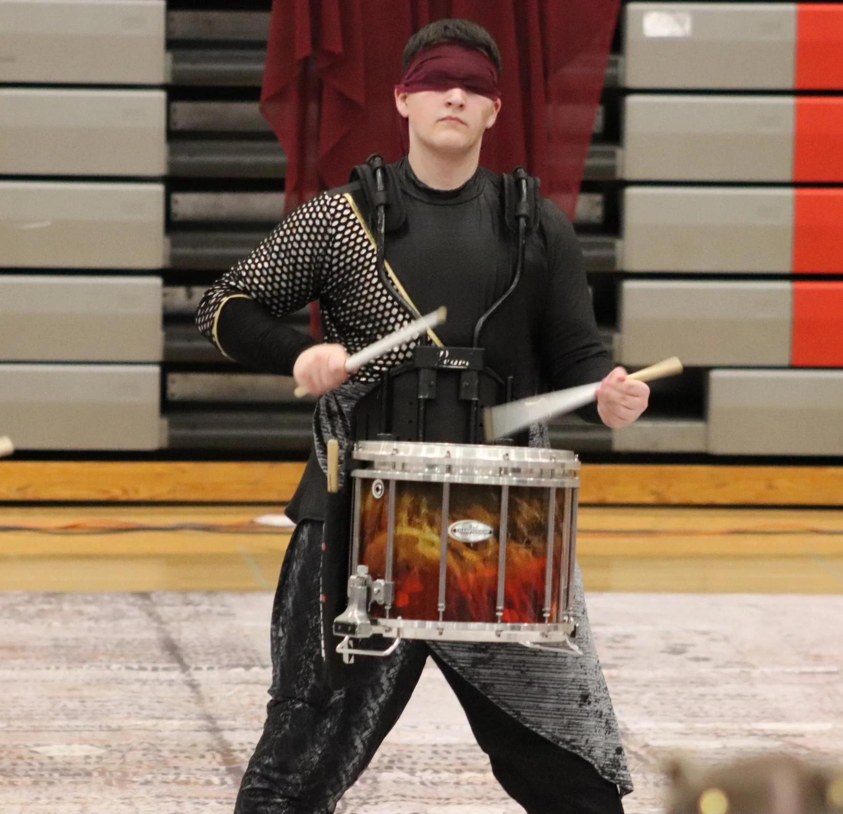 A young man blindfolded, playing a marching snare drum with drumsticks, in a gymnasium for a winter indoor percussion open class competition. 