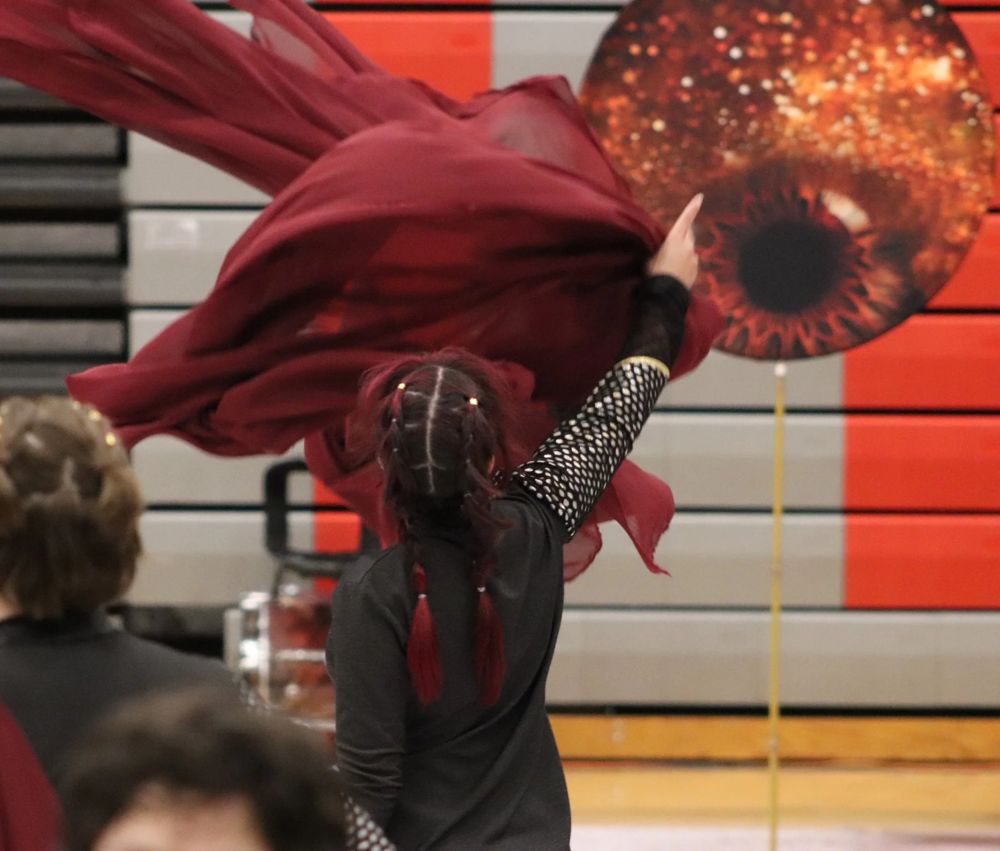 A girl in a black and red outfit is dancing or performing in a winter percussion indoor drumline show. She is reaching towards a large prop in the background that resembles an eye with a fiery orange and red iris. visual effects 