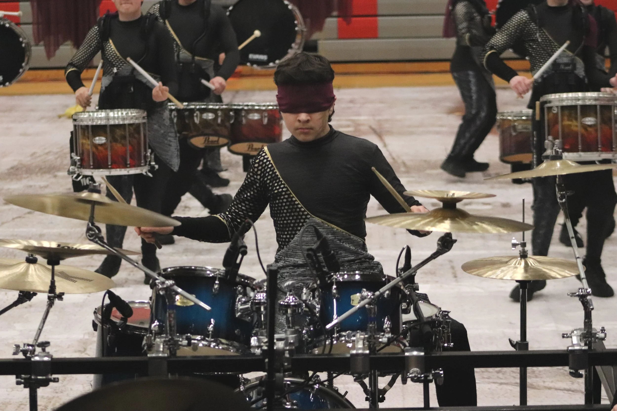 A front ensemble drumline member playing a drum set with other drummers in the background, in an indoor gymnasium for a winter indoor percussion show. 