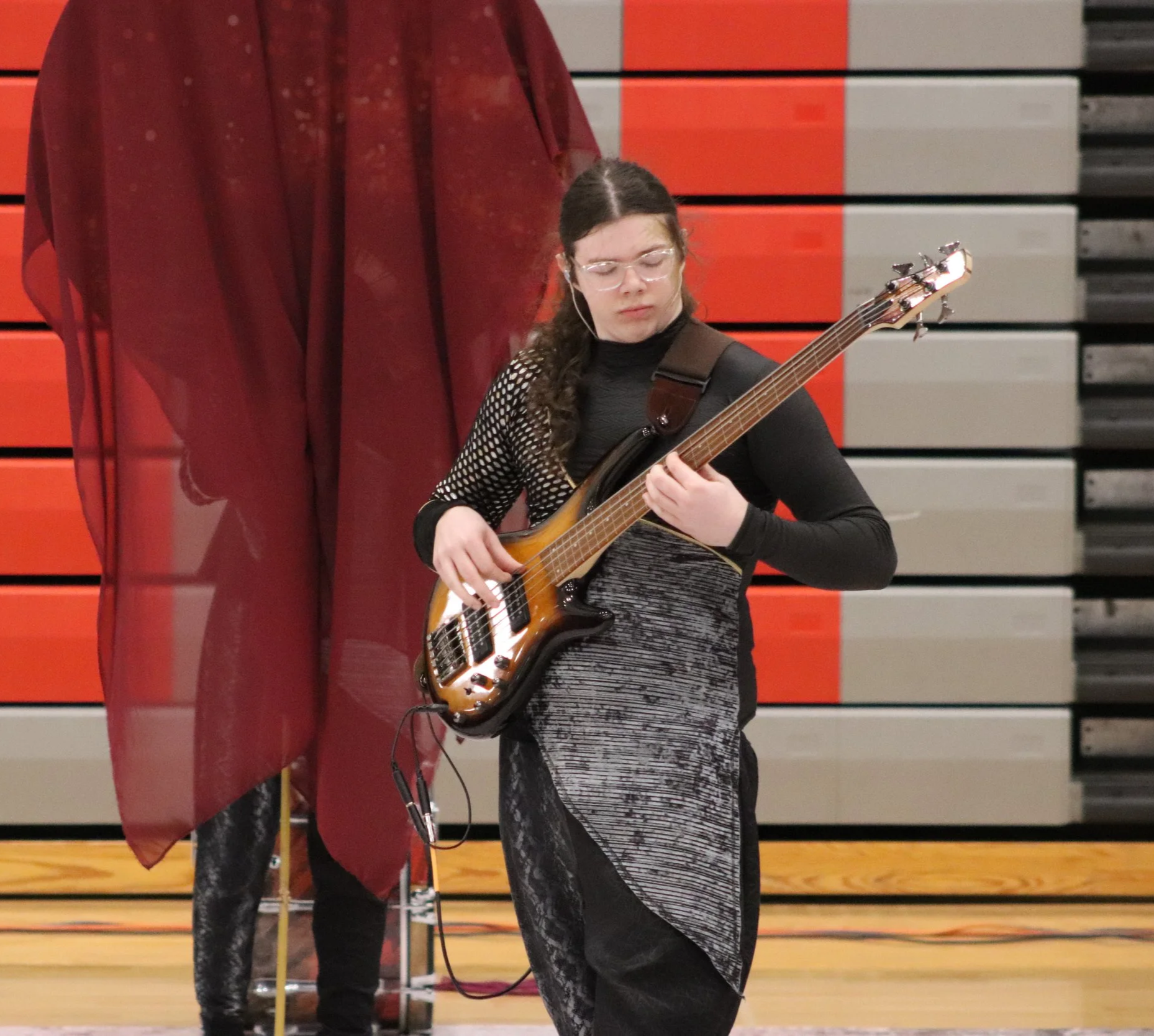 A young man playing an electric bass guitar on a stage with a wooden floor and colorful backdrop in a winter percussion drumline front ensemble show competition.