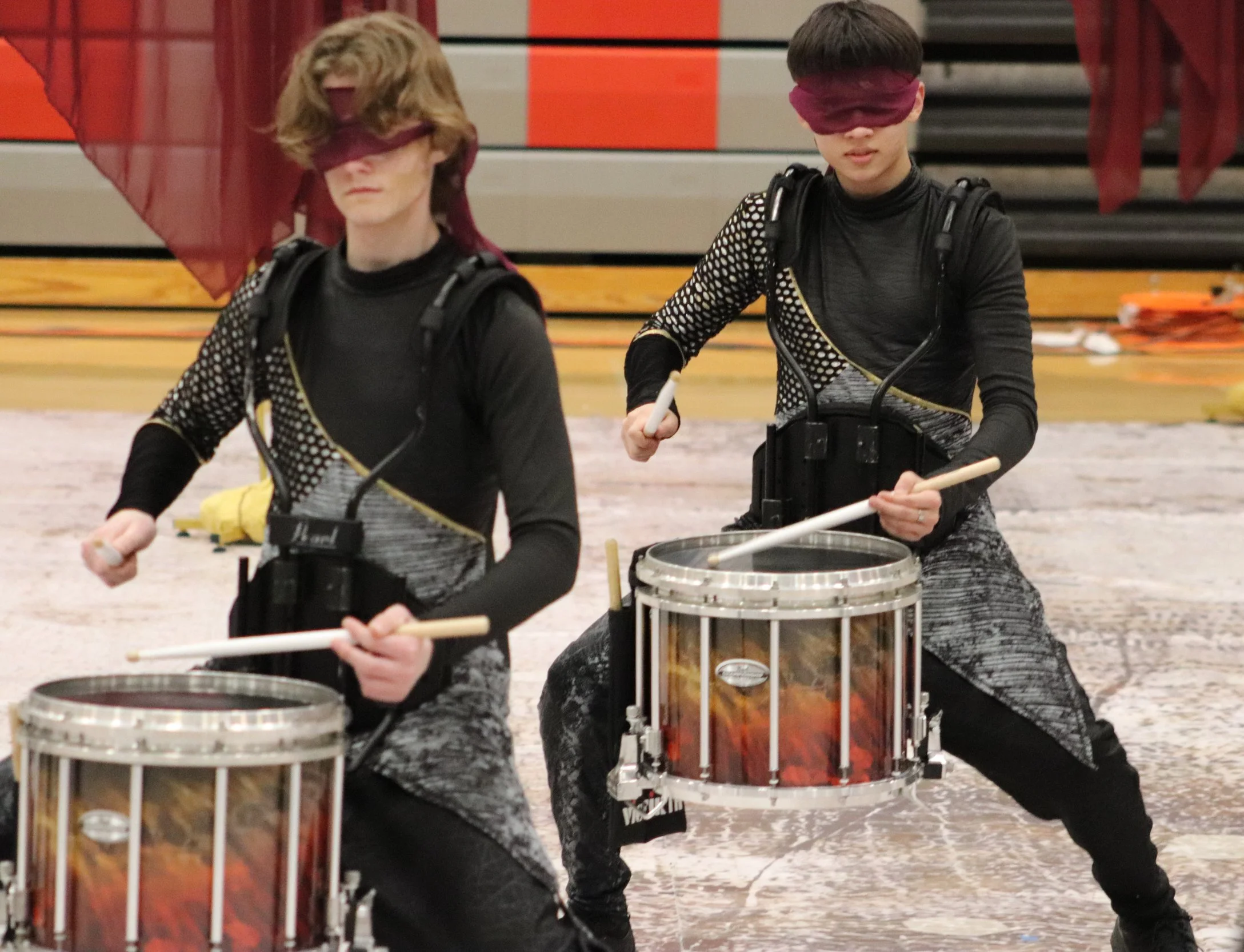 Two young musicians blindfolded, playing marching snare drums on a stage with a gymnasium background, wearing black and silver outfits for a winter indoor percussion open class competition. 