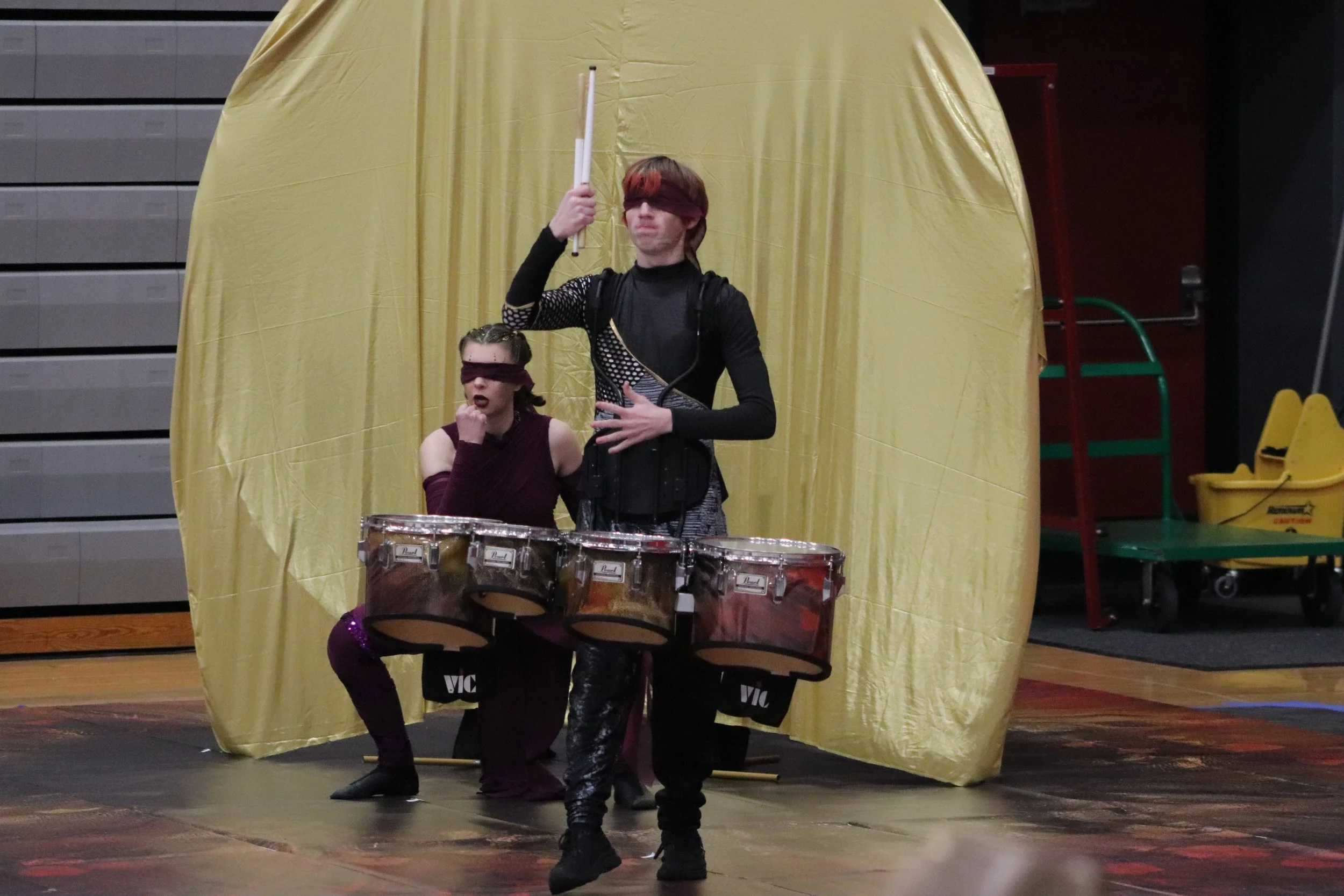 Two performers playing tenor drums with blindfolds, one standing and playing tenor drums with a drumstick, one crouched in movement and dance, in front of a yellow backdrop.