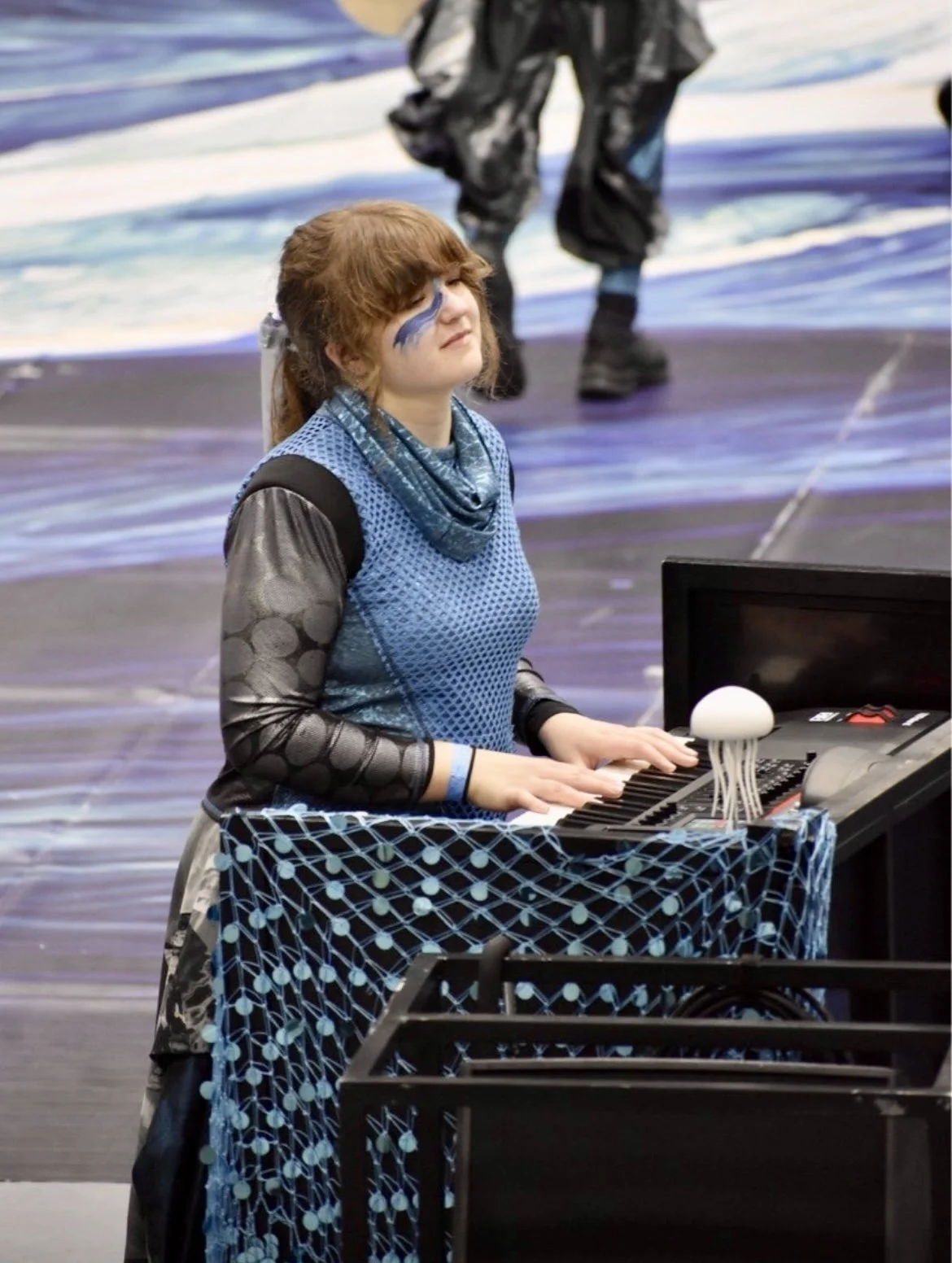 A young woman with reddish-brown hair and face paint playing a keyboard at a winter indoor percussion drumline performance.