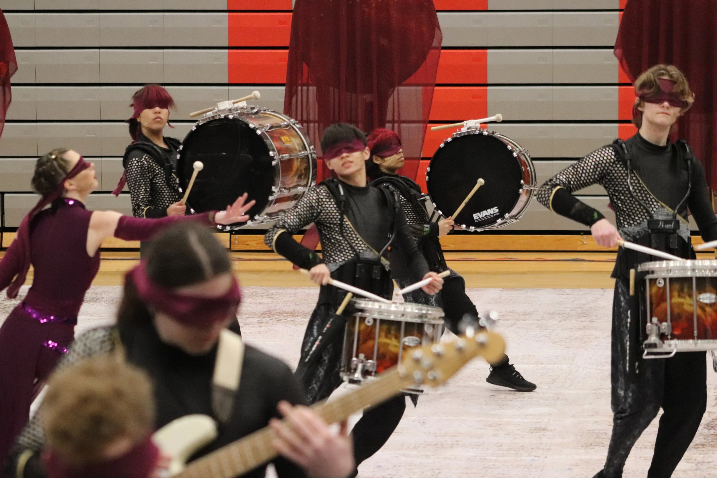 A group of young performers playing drums and guitar during a performance, with some wearing blindfolds, on a gymnasium floor with a decorated backdrop for a winter indoor percussion drumline show. 
