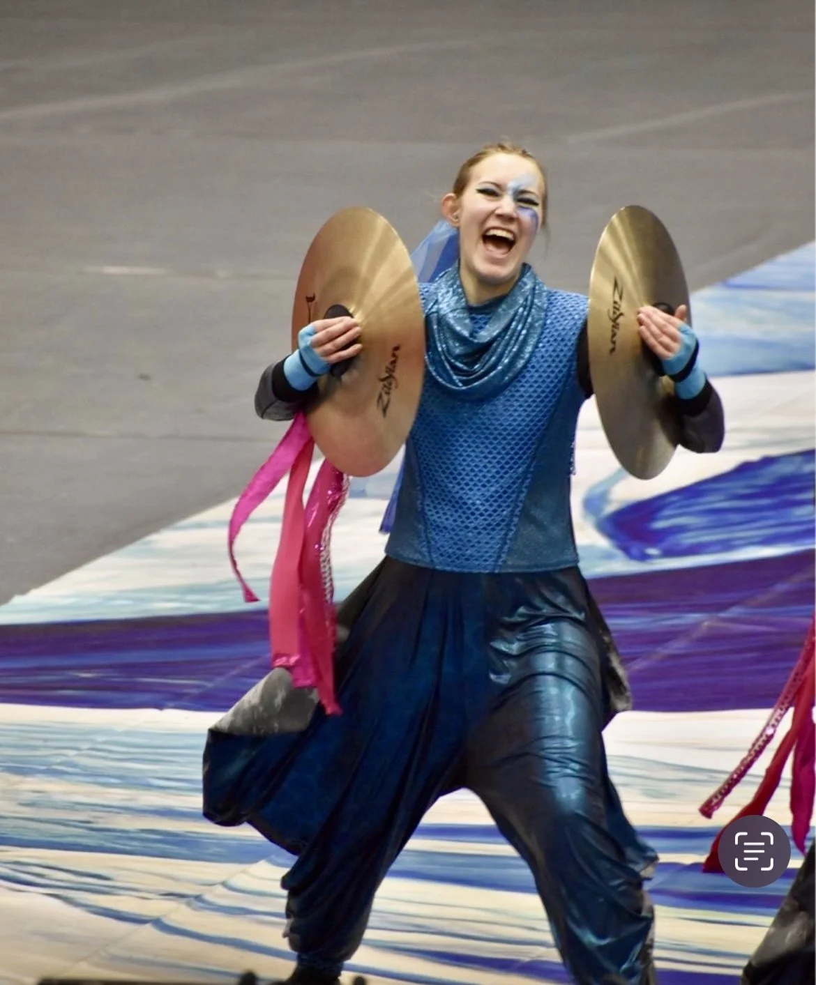 A performer holding cymbals on stage, smiling and wearing colorful performance attire as part of a winter indoor percussion drumline performance.