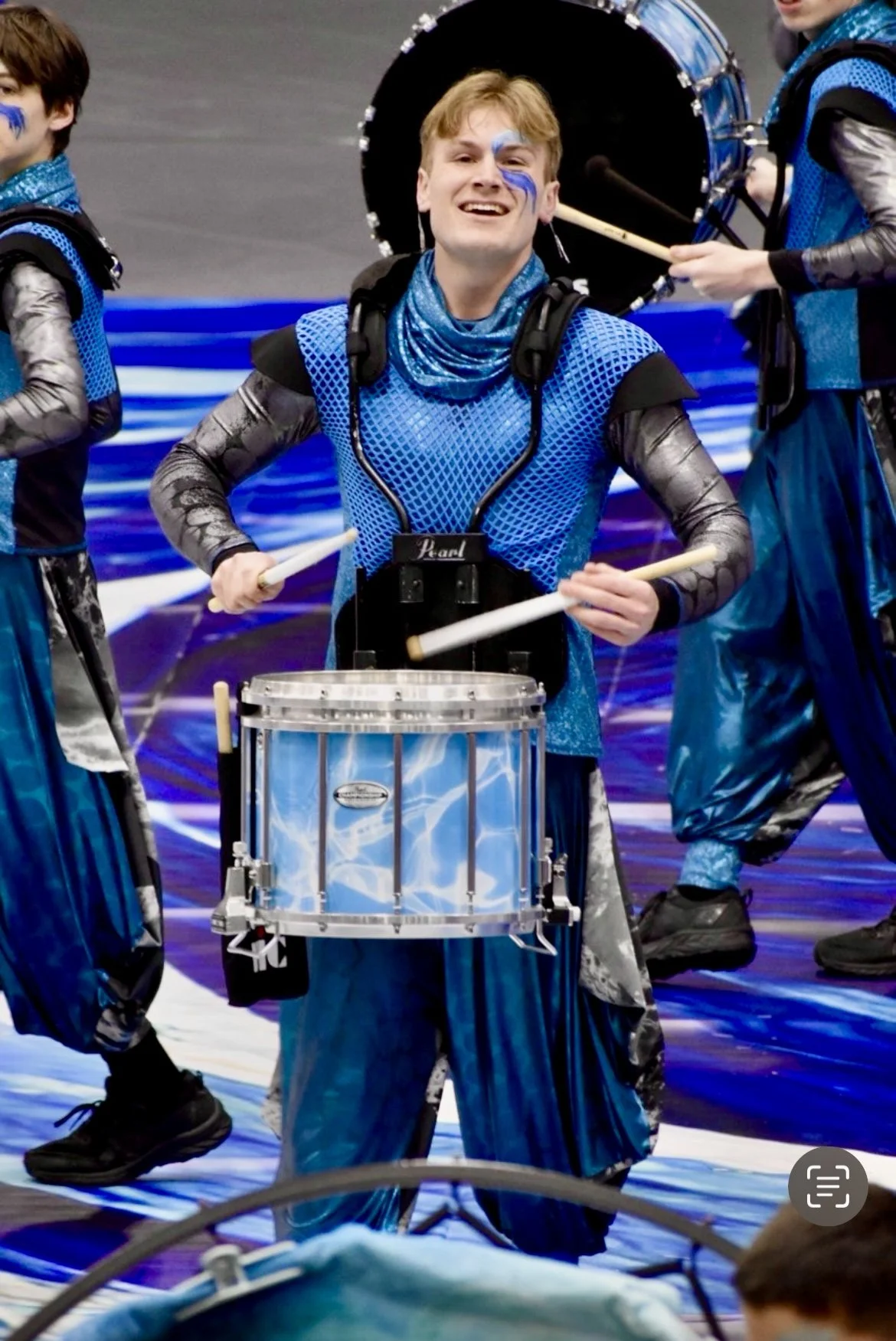 A young man smiling and playing a snare drum while dressed in a blue and black costume with face paint, part of a winter indoor percussion drumline performance.